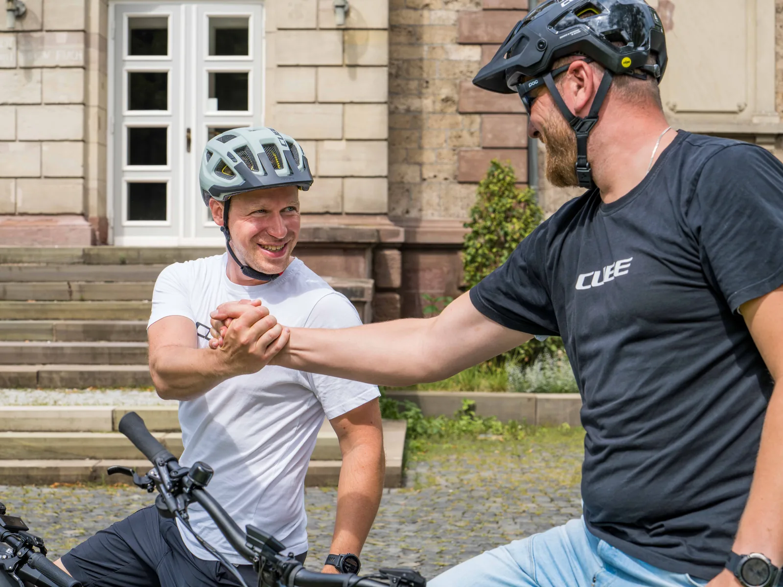 Zwei Radfahrer begrüßen sich per Handschlag vor einer Steintreppe, einer im weißen Shirt auf dem Rad, der andere im schwarzen CUBE-Shirt daneben.