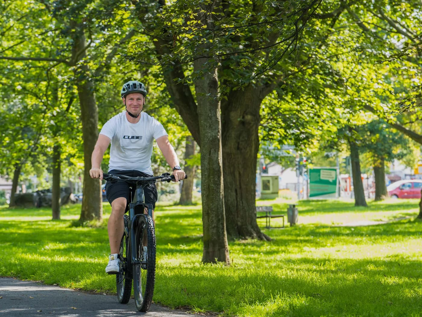 Ein Radfahrer im weißen CUBE-Shirt fährt durch einen sonnigen Park mit alten Bäumen auf die Kamera zu.