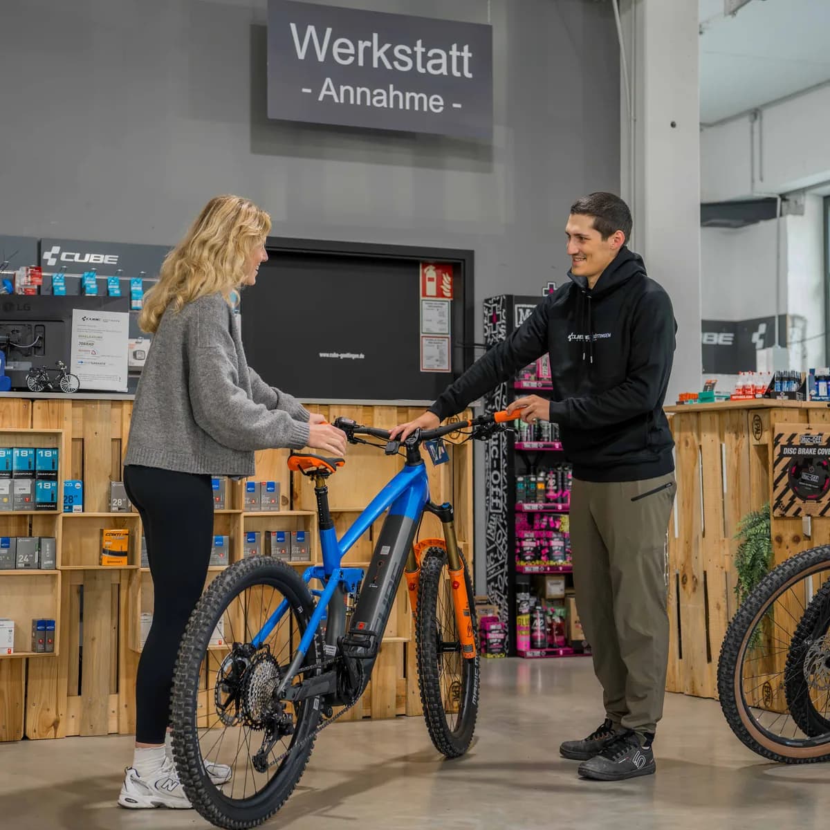 Beratung an der Werkstattannahme im CUBE Store Göttingen: eine Kundin und ein Mitarbeiter stehen links und rechts eines blau-orangen Mountainbikes.