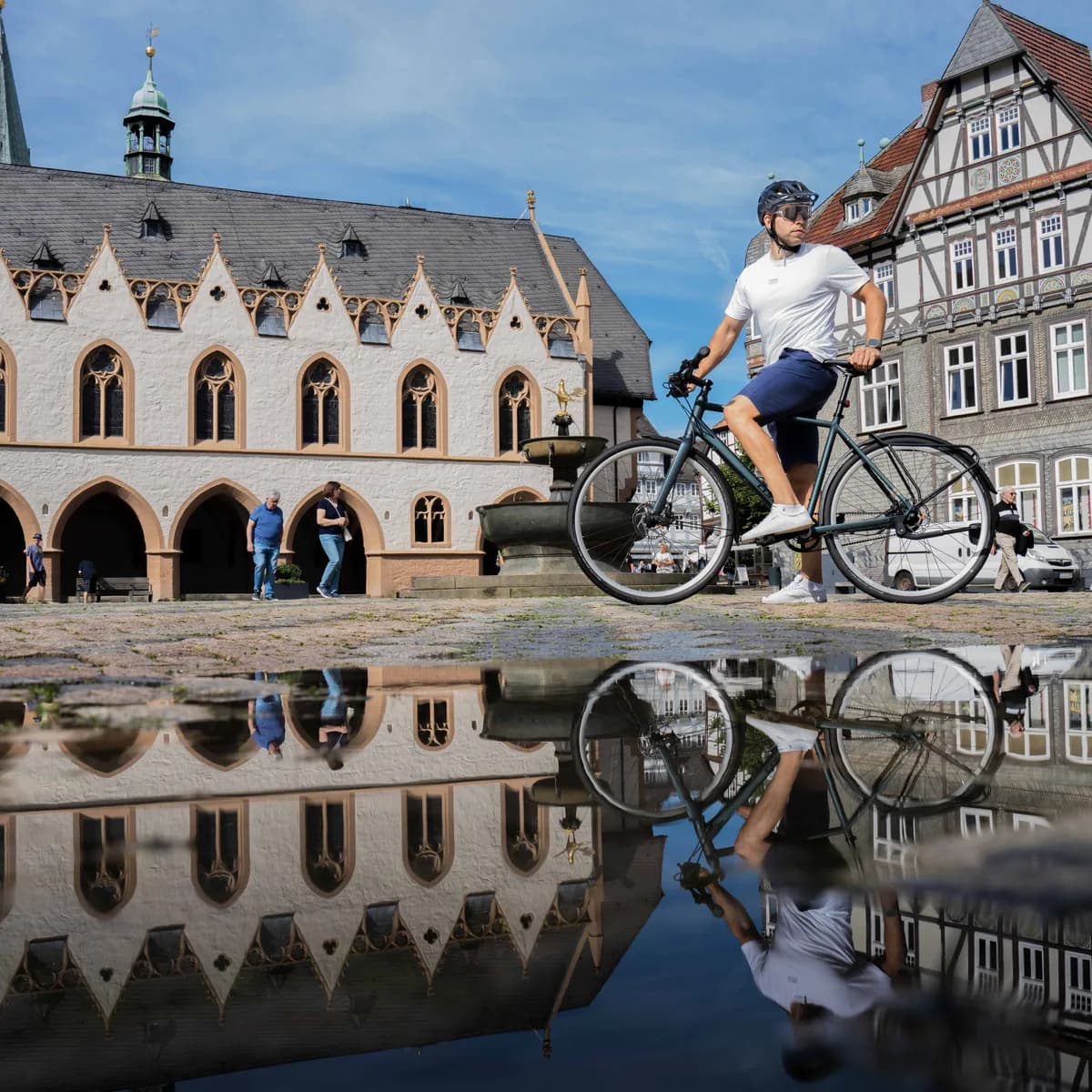 Ein Radfahrer auf dem Marktplatz von Goslar, davor eine Pfütze, in der sich das historische Rathaus und die Fachwerkhäuser spiegeln.