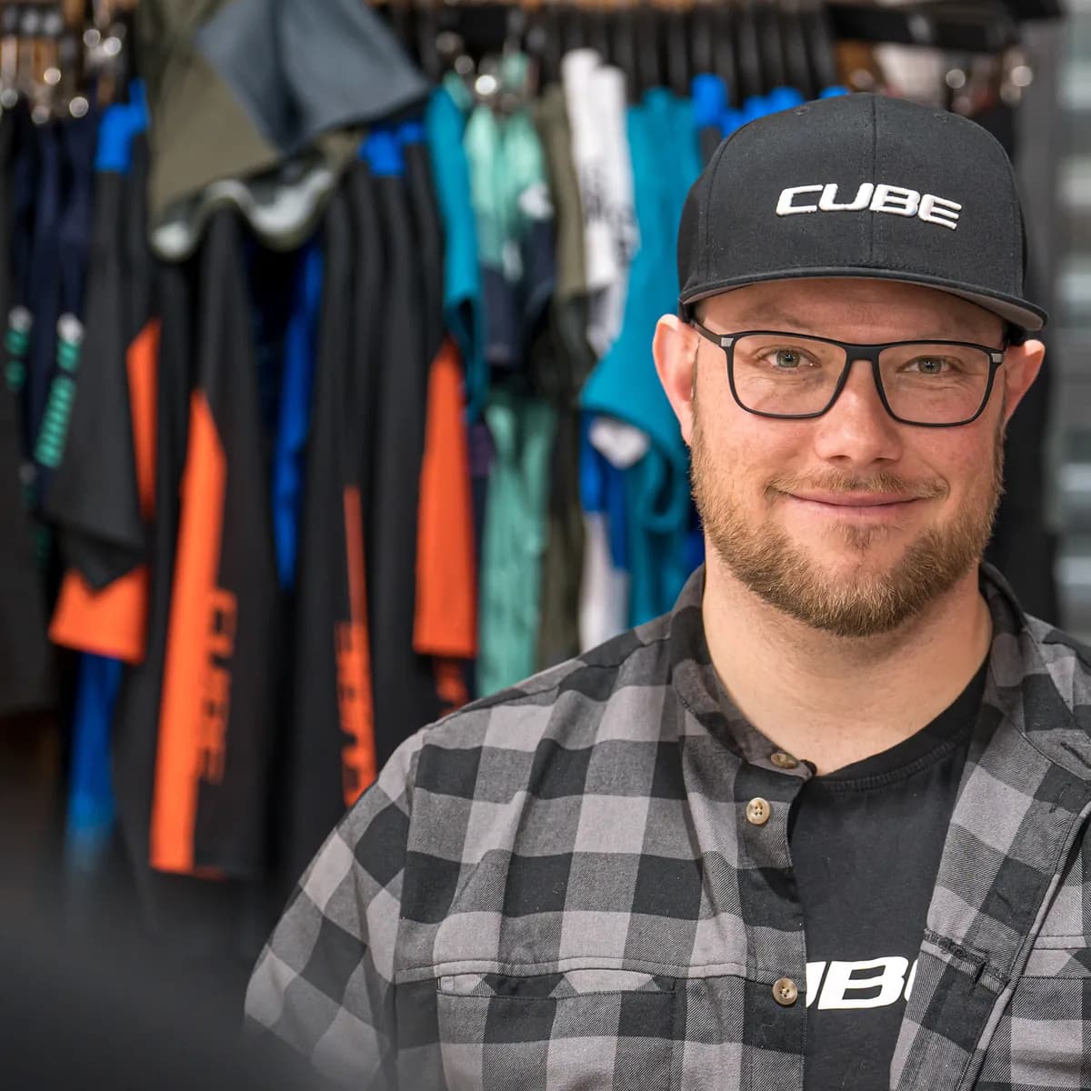 Mitarbeiter des CUBE Store Hildesheim mit CUBE-Cap und Brille, hinter ihm eine Stange mit Radbekleidung.