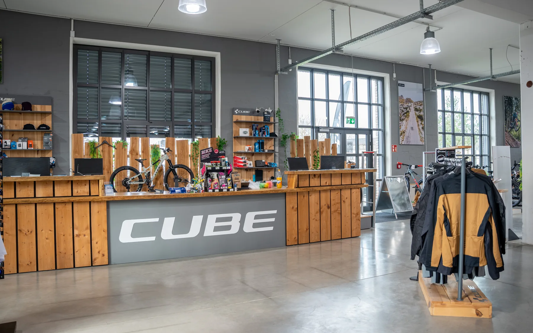 Der Verkaufsraum des CUBE Store Göttingen: ein langer Holztresen mit dem CUBE-Schriftzug, dahinter Arbeitsplätze und Regale, rechts Bekleidung auf einem Ständer.