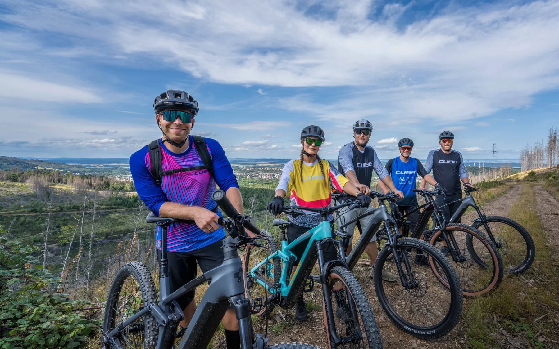 Fünf Radfahrerinnen und Radfahrer in bunten Trikots stehen mit ihren Mountainbikes auf einem Waldweg über Goslar, hinter ihnen die Landschaft des Harzes.