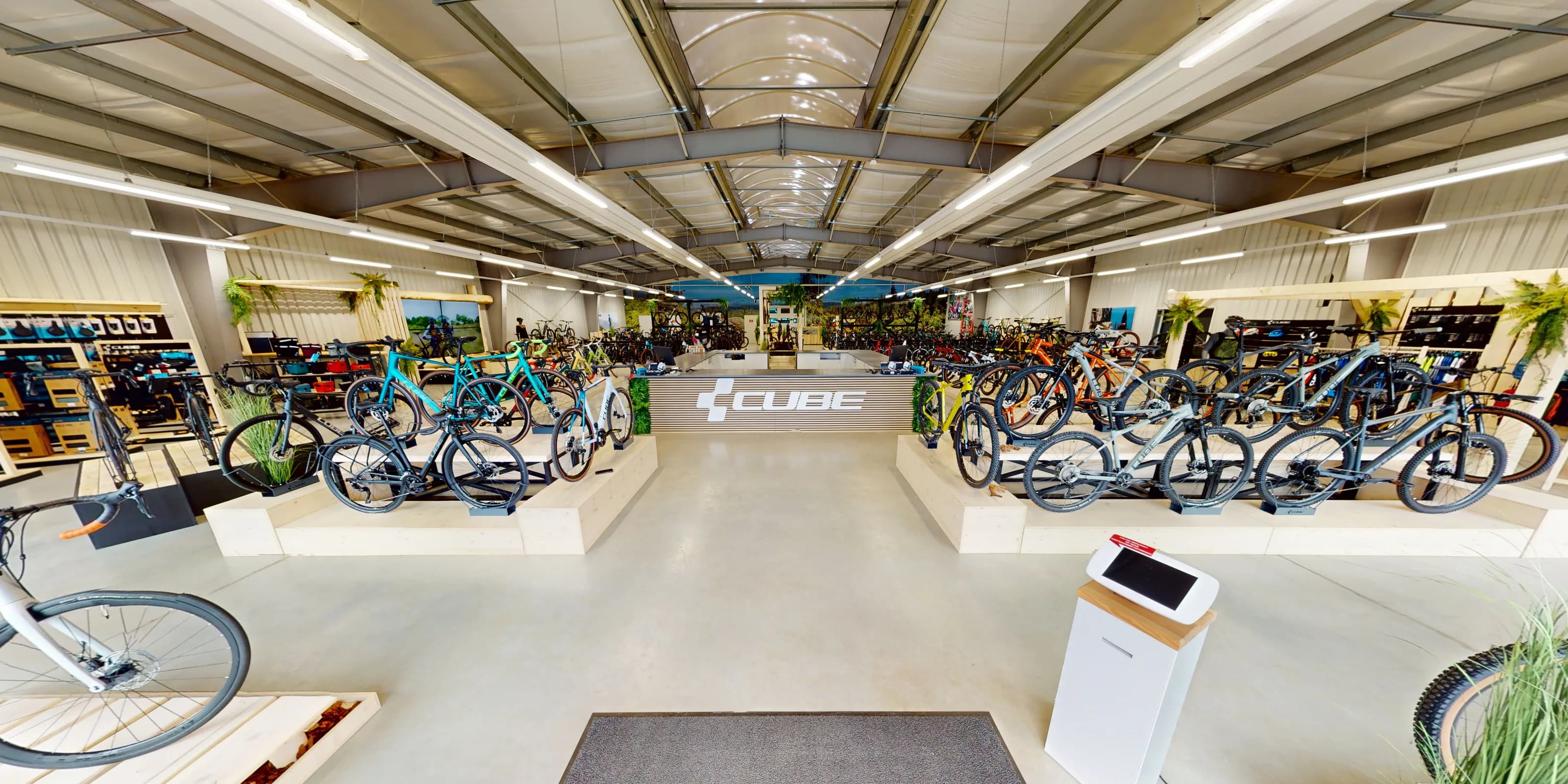 Virtueller Rundgang für den CUBE Store Goslar: der Blick von der Eingangsmatte den Mittelgang hinunter auf die Verkaufstheke mit dem CUBE-Schriftzug, links und rechts Räder auf den Holzpodesten.