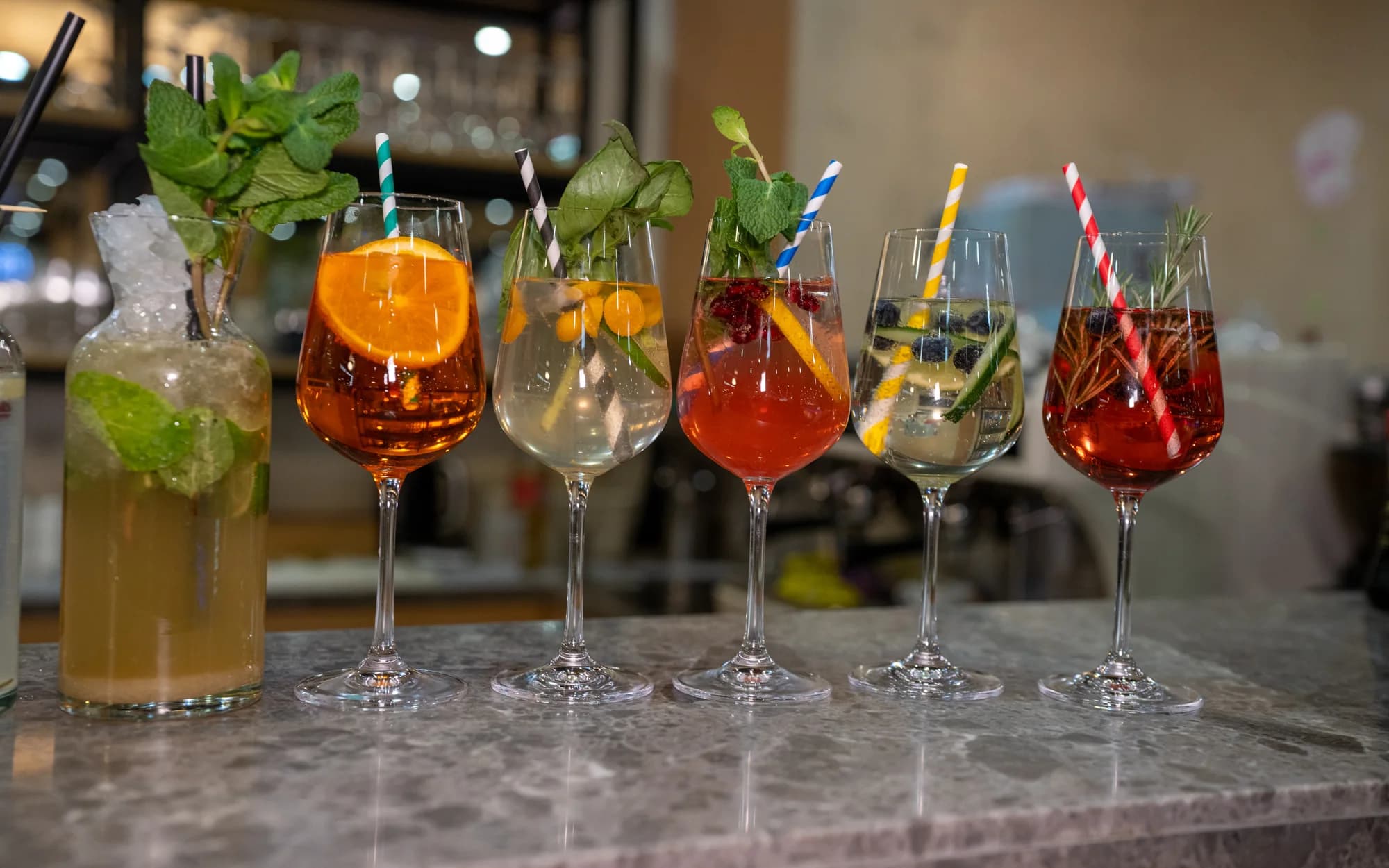 Sechs Cocktails in Longdrink- und Weingläsern mit Minze, Orangenscheiben und bunten Strohhalmen stehen nebeneinander auf der Marmortheke der Bar.