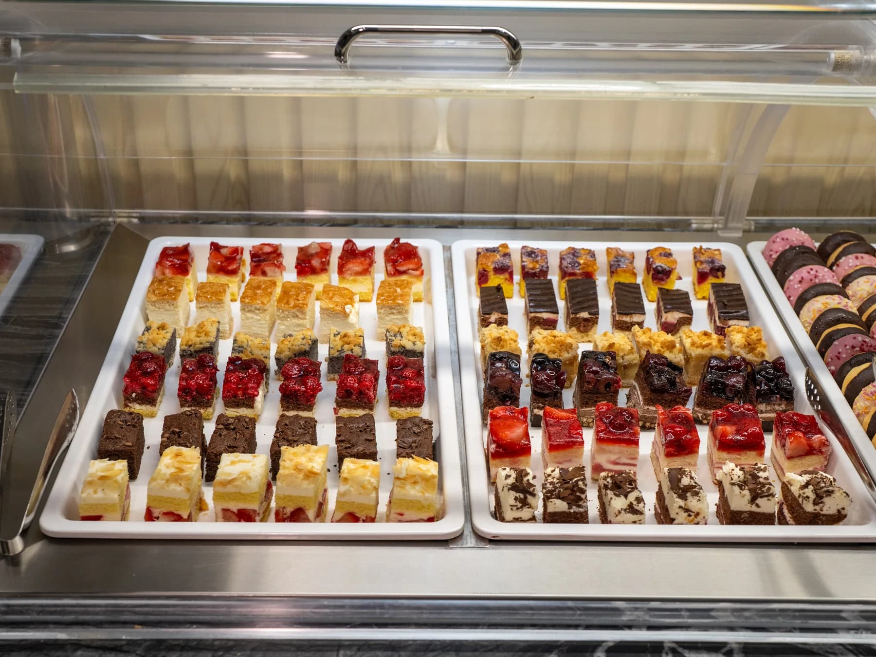 Zwei Bleche mit Dessertschnitten in Reihen, Fruchtspiegel und Schokolade, in der gekühlten Vitrine des Buffets.