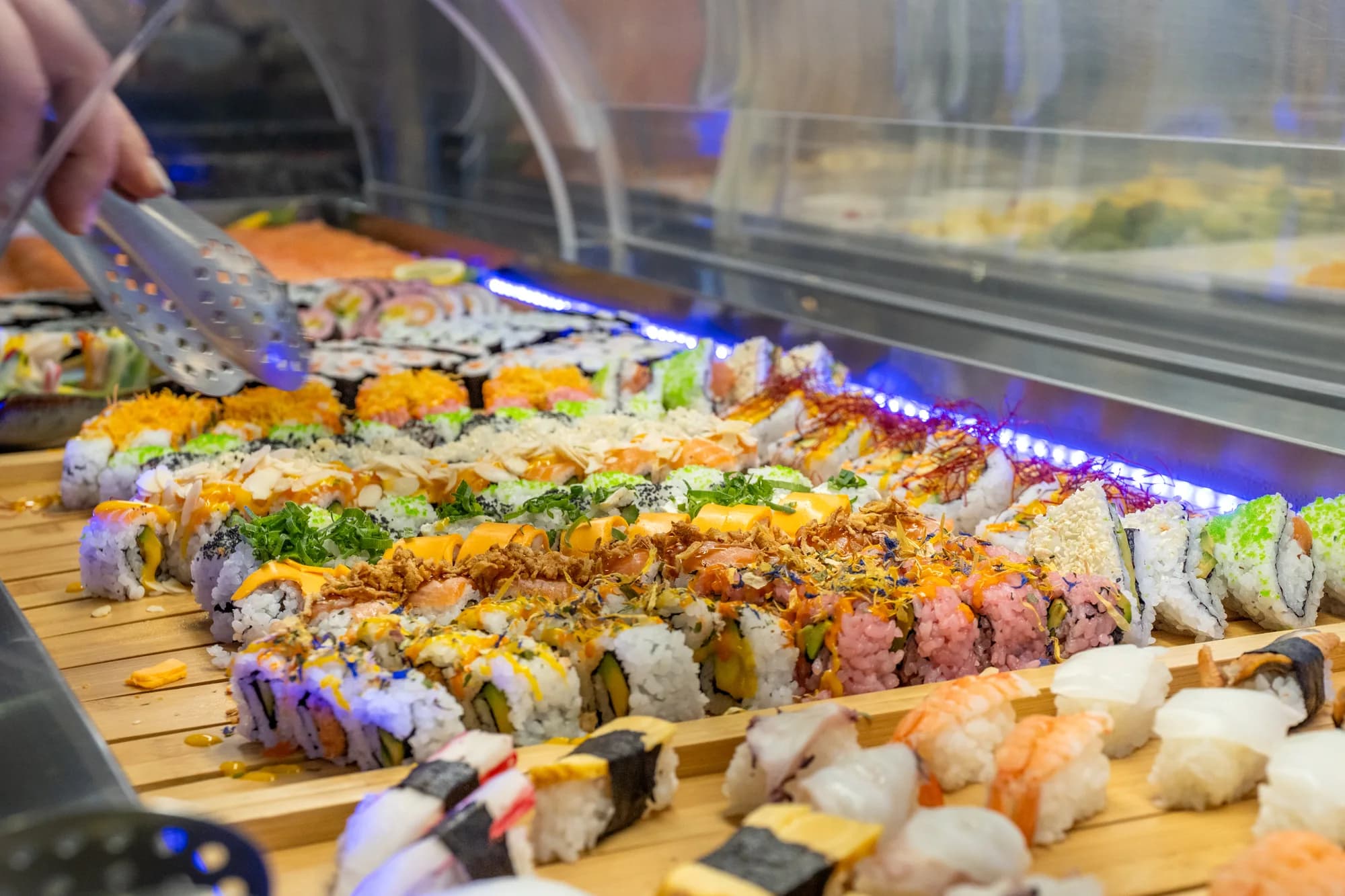 Eine Hand hebt mit der Zange eine Maki-Rolle von der Sushitheke; die Reihen laufen unter dem blauen Lichtband und der Vitrinenhaube in die Tiefe.