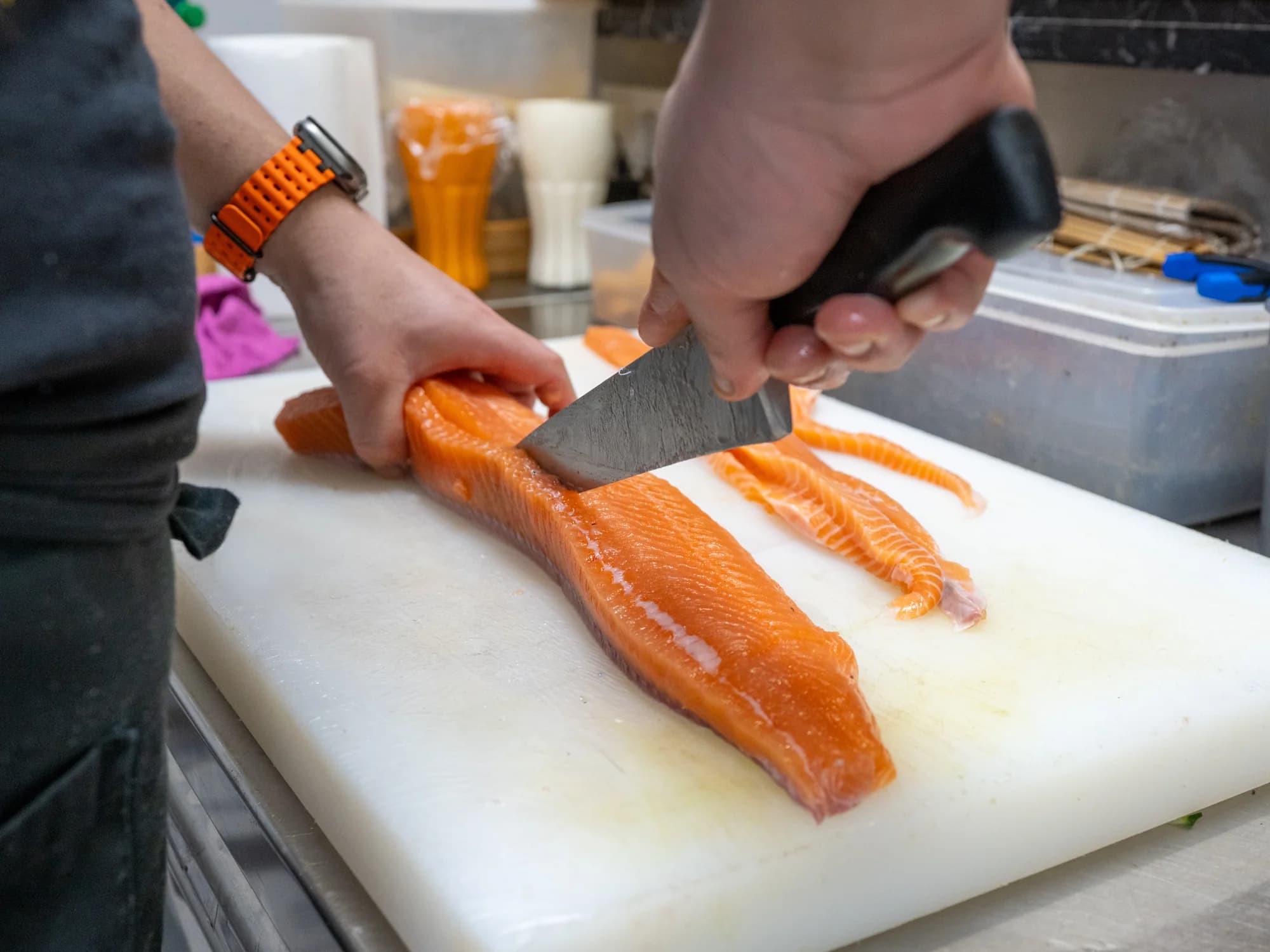Zwei Hände schneiden mit einem langen Messer Streifen von einem Lachsfilet, das auf einem weissen Schneidebrett in der Küche liegt.