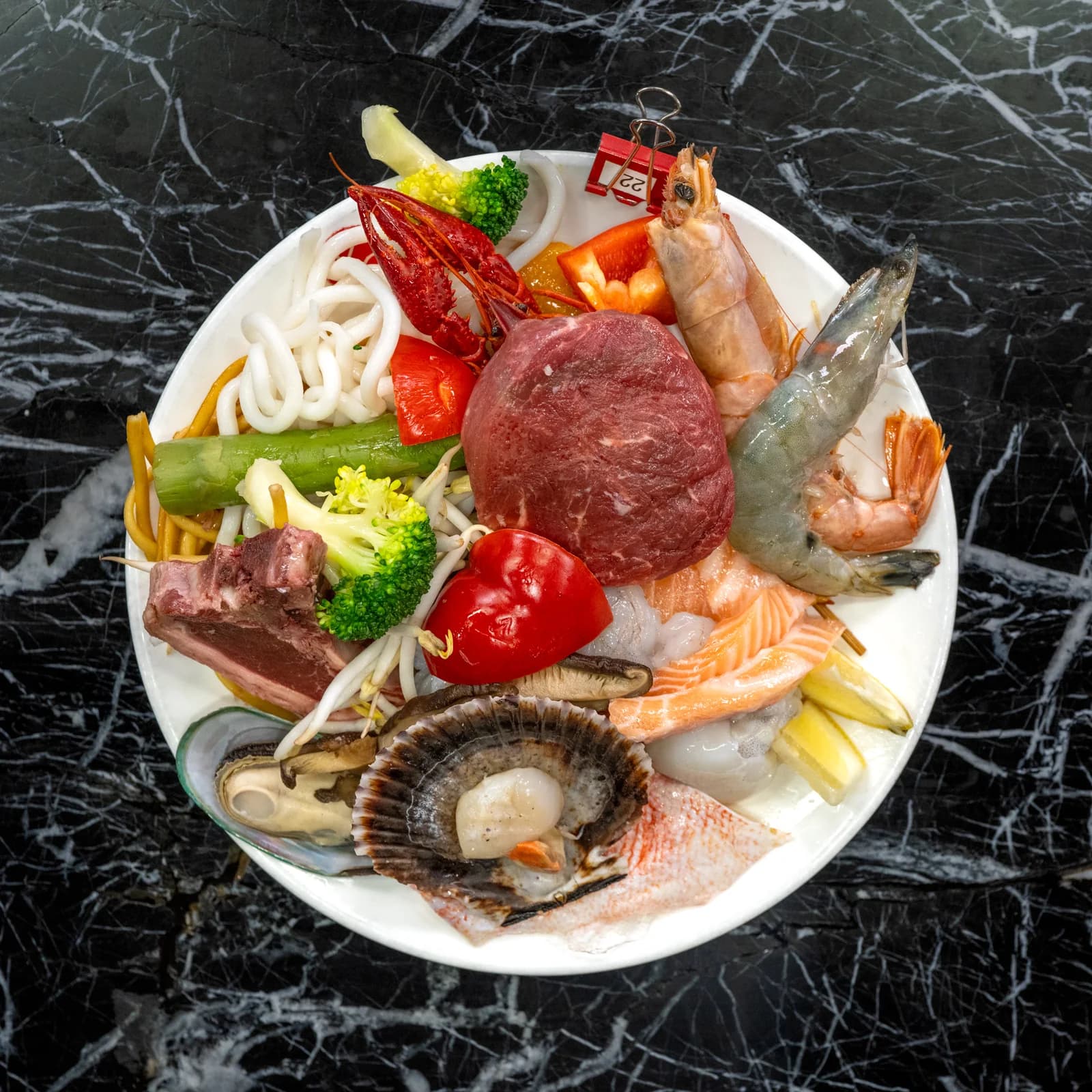 Von oben: ein Teller mit Garnelen, Jakobsmuschel, Tintenfisch, Nudeln und Paprika auf schwarzer Marmorfläche.
