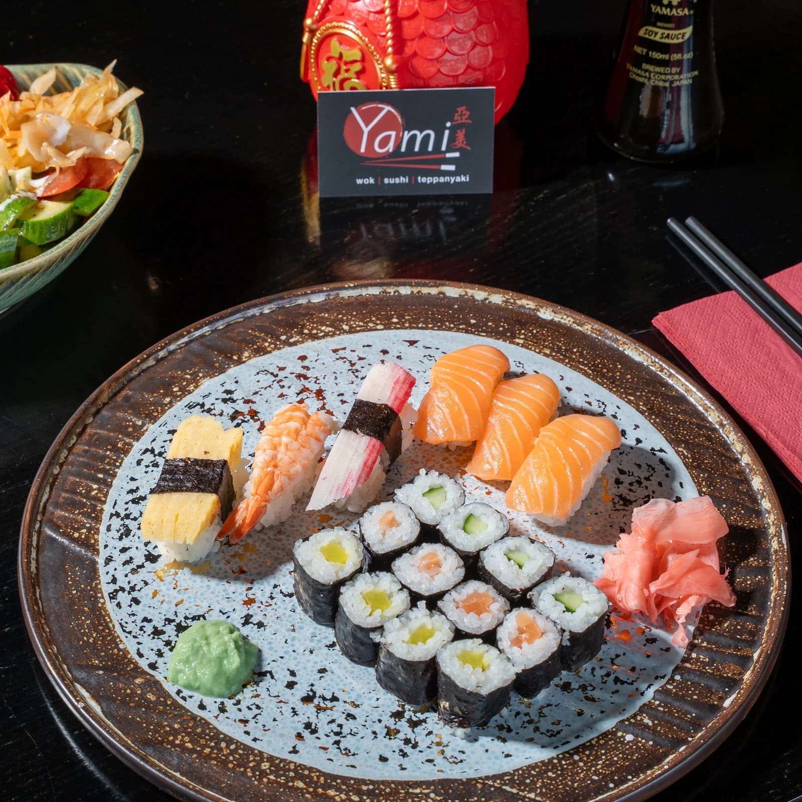Eine runde Steinplatte mit Nigiri, Maki und Inside-out-Rollen, daneben Wasabi und eingelegter Ingwer, im Hintergrund die rote Tischkarte des Lokals.