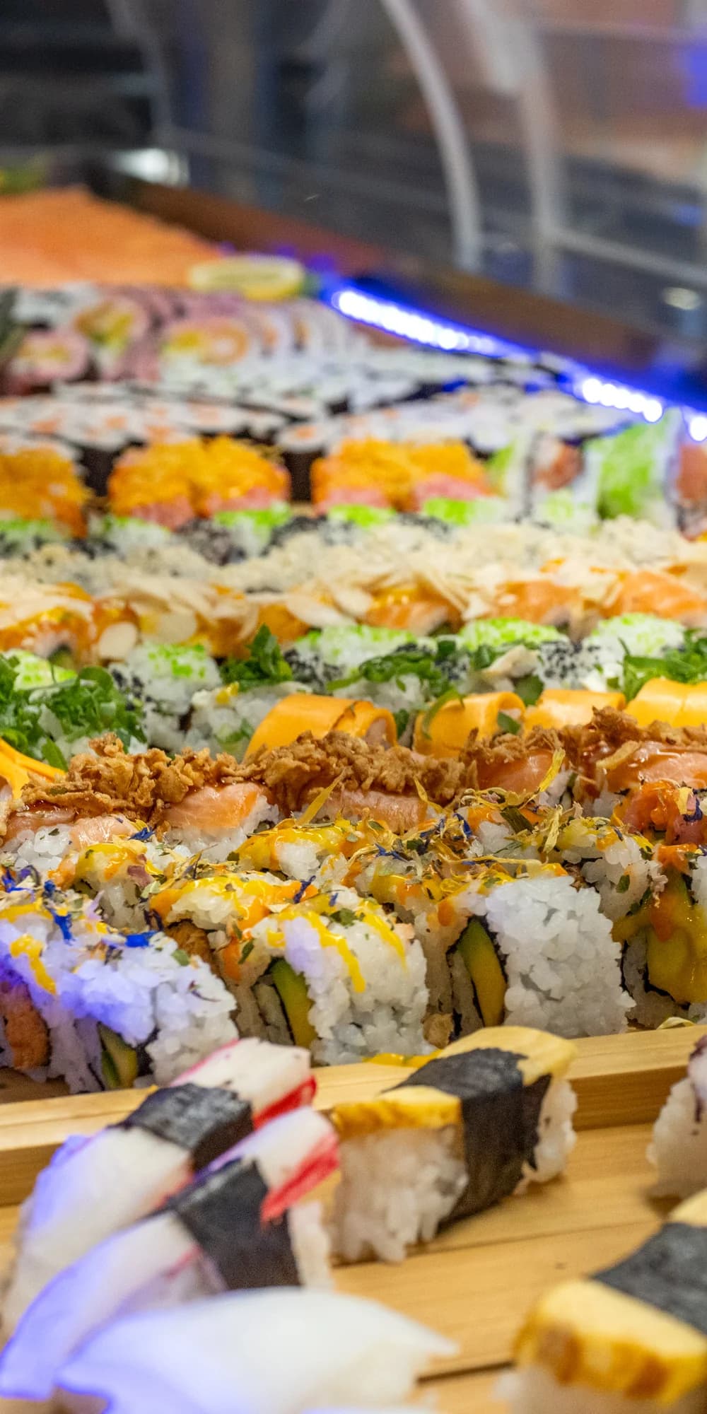 Eine lange Buffettheke mit Maki-Rollen und Nigiri auf Holzbrettern, darüber ein blaues Lichtband, die Reihe läuft in die Tiefe des Raums.