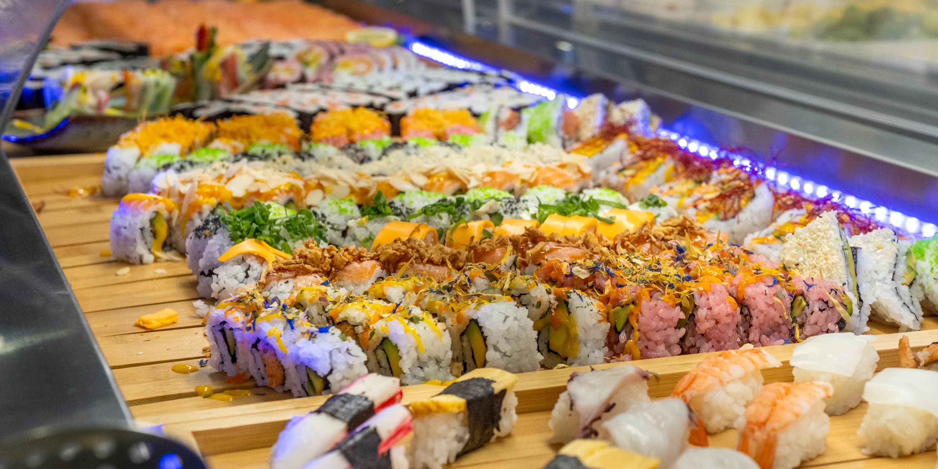Asia Restaurant YAMI: Eine lange Buffettheke mit Maki-Rollen und Nigiri auf Holzbrettern, darüber ein blaues Lichtband, die Reihe läuft in die Tiefe des Raums.