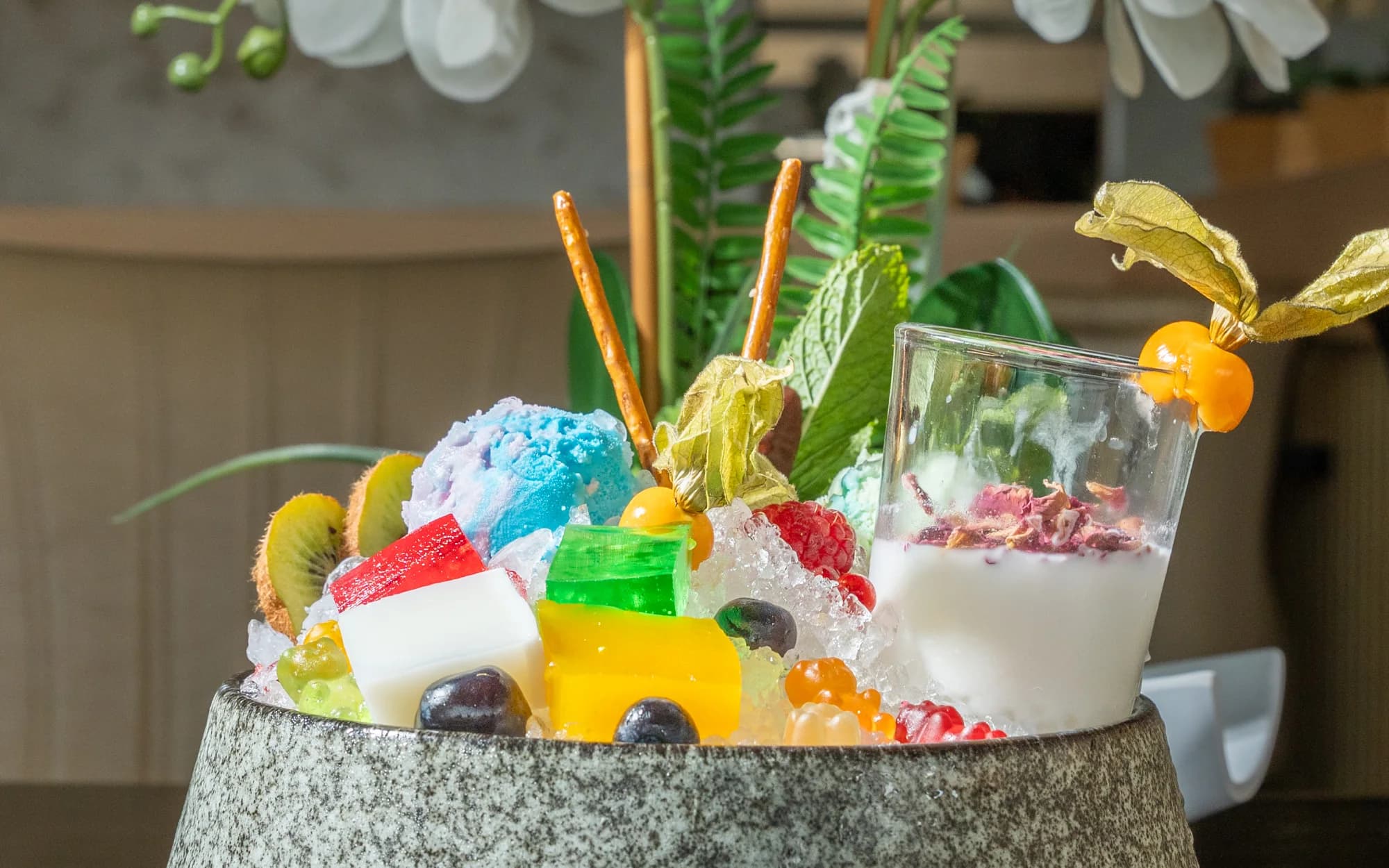 Ein Steintopf mit geschabtem Eis, buntem Gelee, Kiwi, Beeren und einer Physalis, daneben ein Glas mit Joghurt und Blüten.