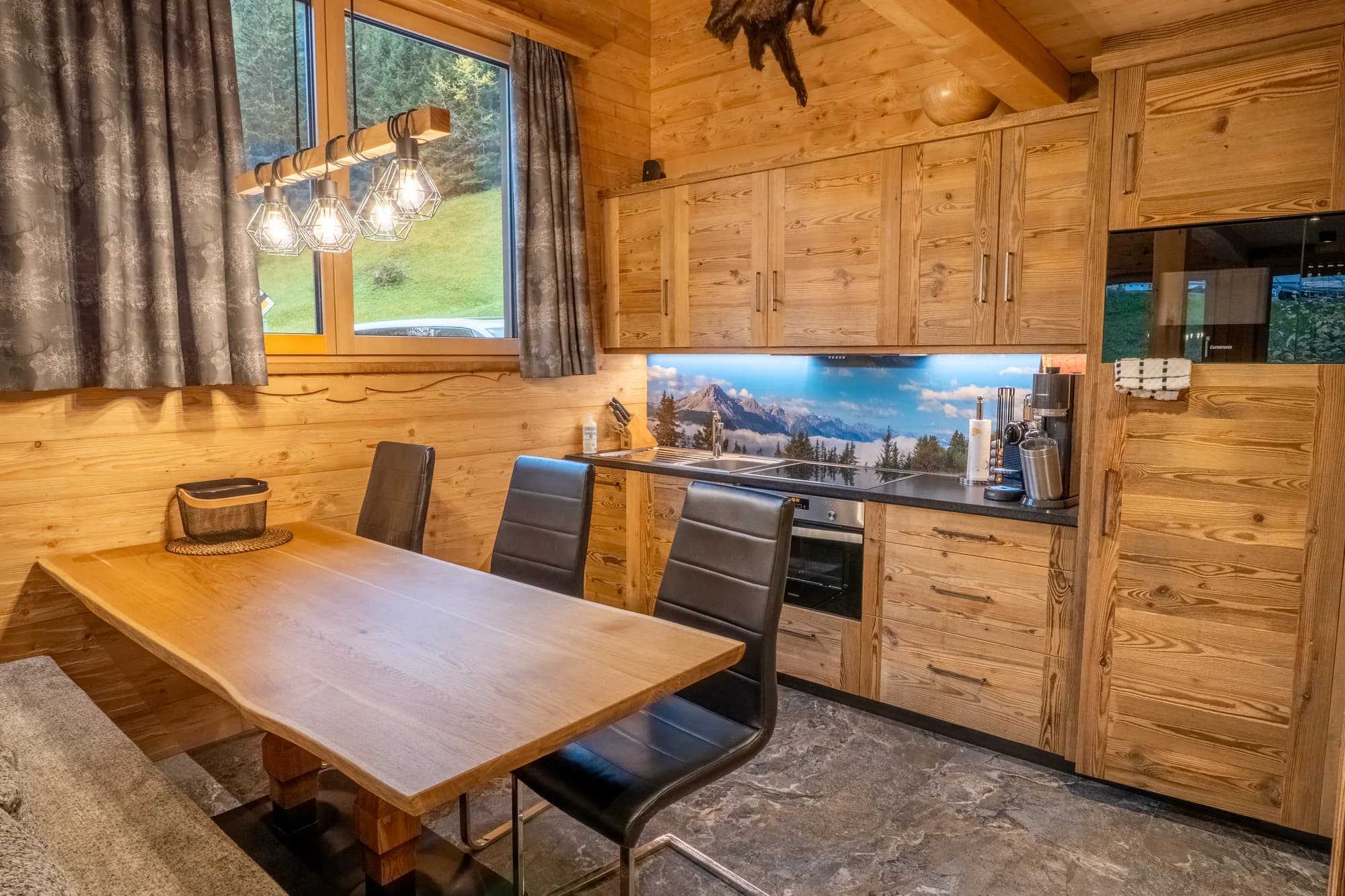 Der Essplatz im Blockhaus Fuchsbau: ein langer Holztisch mit Bank und Stühlen unter einer Reihe kleiner Hängeleuchten, dahinter die Küchenzeile aus Holz.