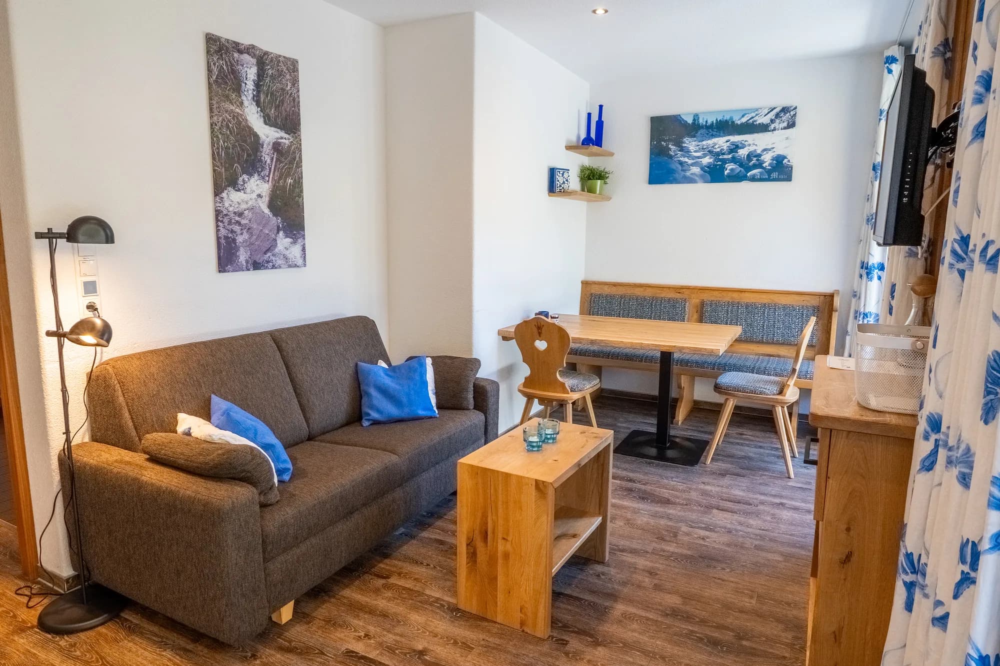 Der Wohnraum der Wohnung Bachrauschen: ein graues Sofa mit blauen Kissen, davor ein Holzwürfel als Tisch, dahinter der Essplatz am Fenster.