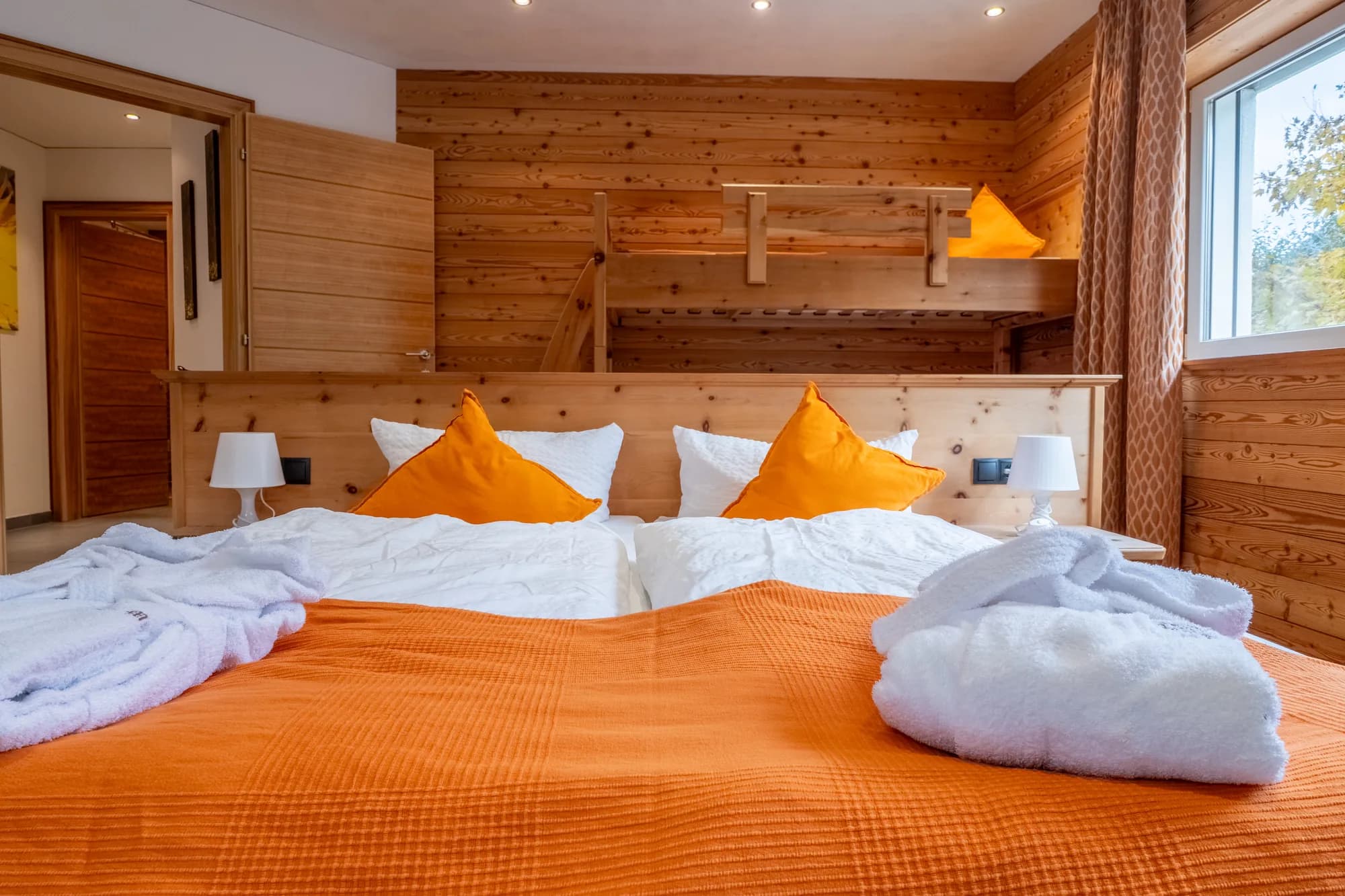 Ein Schlafzimmer der Wohnung Wildwechsel: ein Doppelbett mit oranger Tagesdecke vor einer Zirbenholzwand, rechts ein Stockbett, links das Fenster.