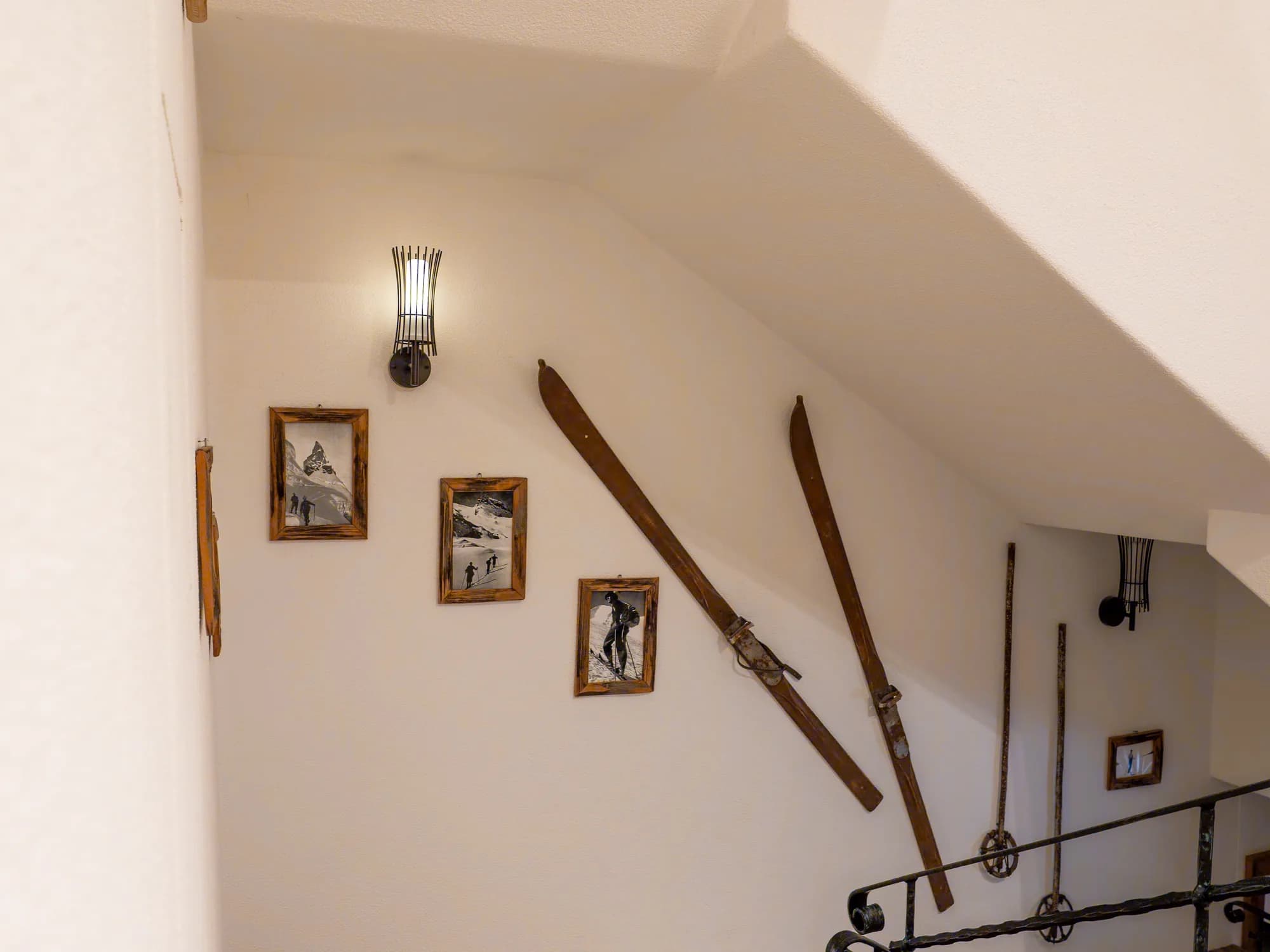 Das Stiegenhaus der Alten Mühle: zwei historische Holzskier und gerahmte schwarzweiße Aufnahmen an der weißen Wand, daneben das schmiedeeiserne Geländer.