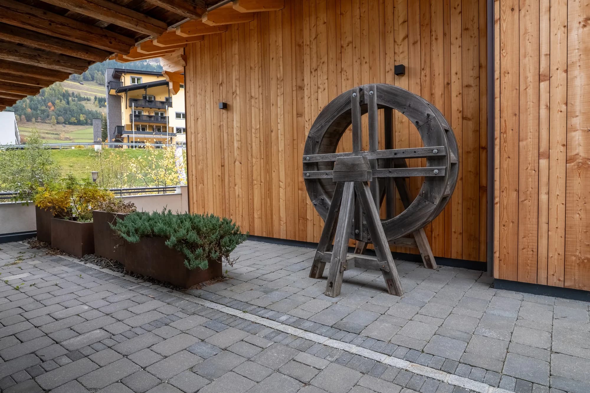 Ein historisches hölzernes Mühlrad an der Holzwand der Alten Mühle, davor zwei Pflanztröge auf dem gepflasterten Vorplatz.