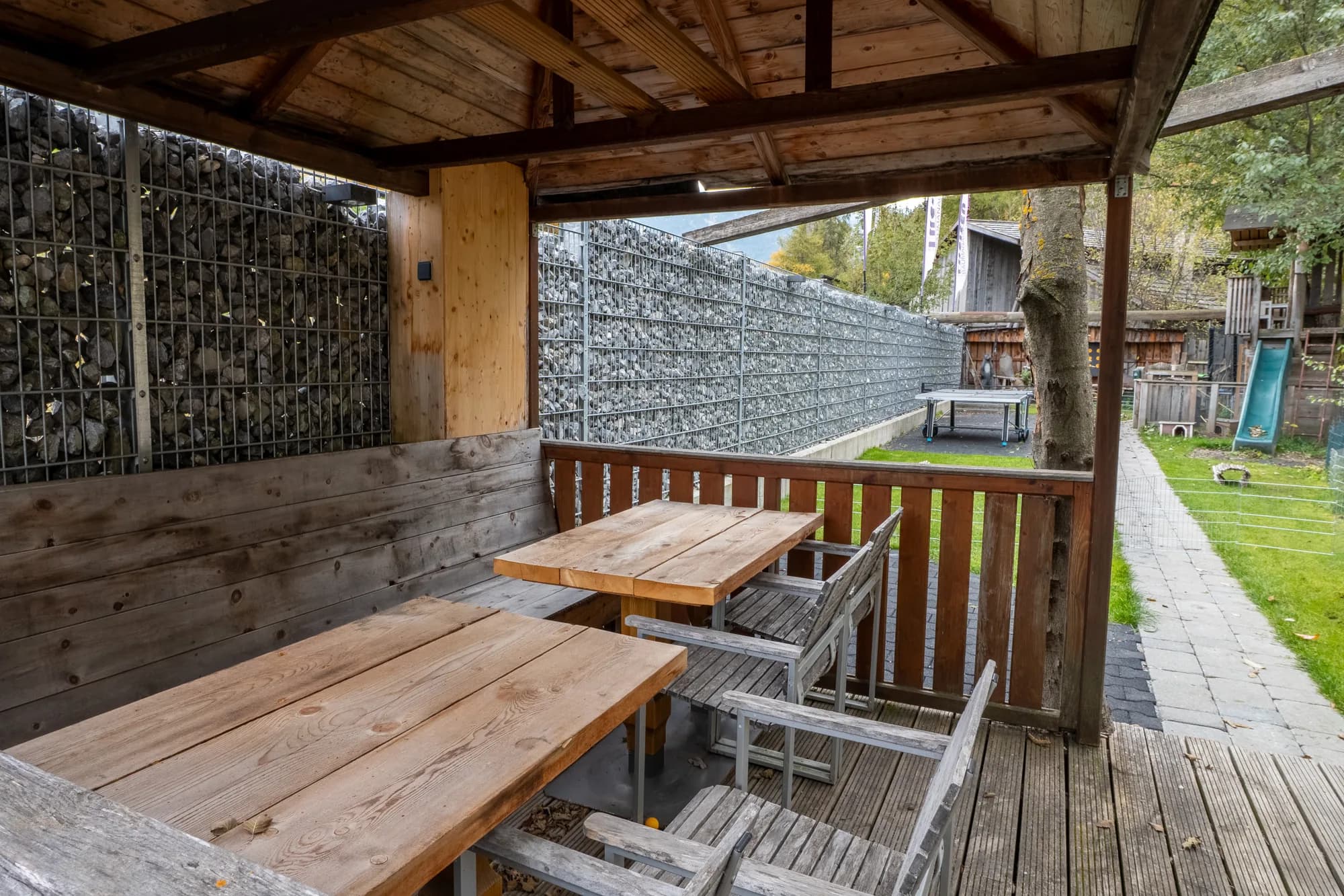 Die gemeinsame Terrasse der Alten Mühle: zwei lange Holztische mit Bänken unter einem offenen Holzdach, dahinter der Garten und eine Wand aus Steinkörben.