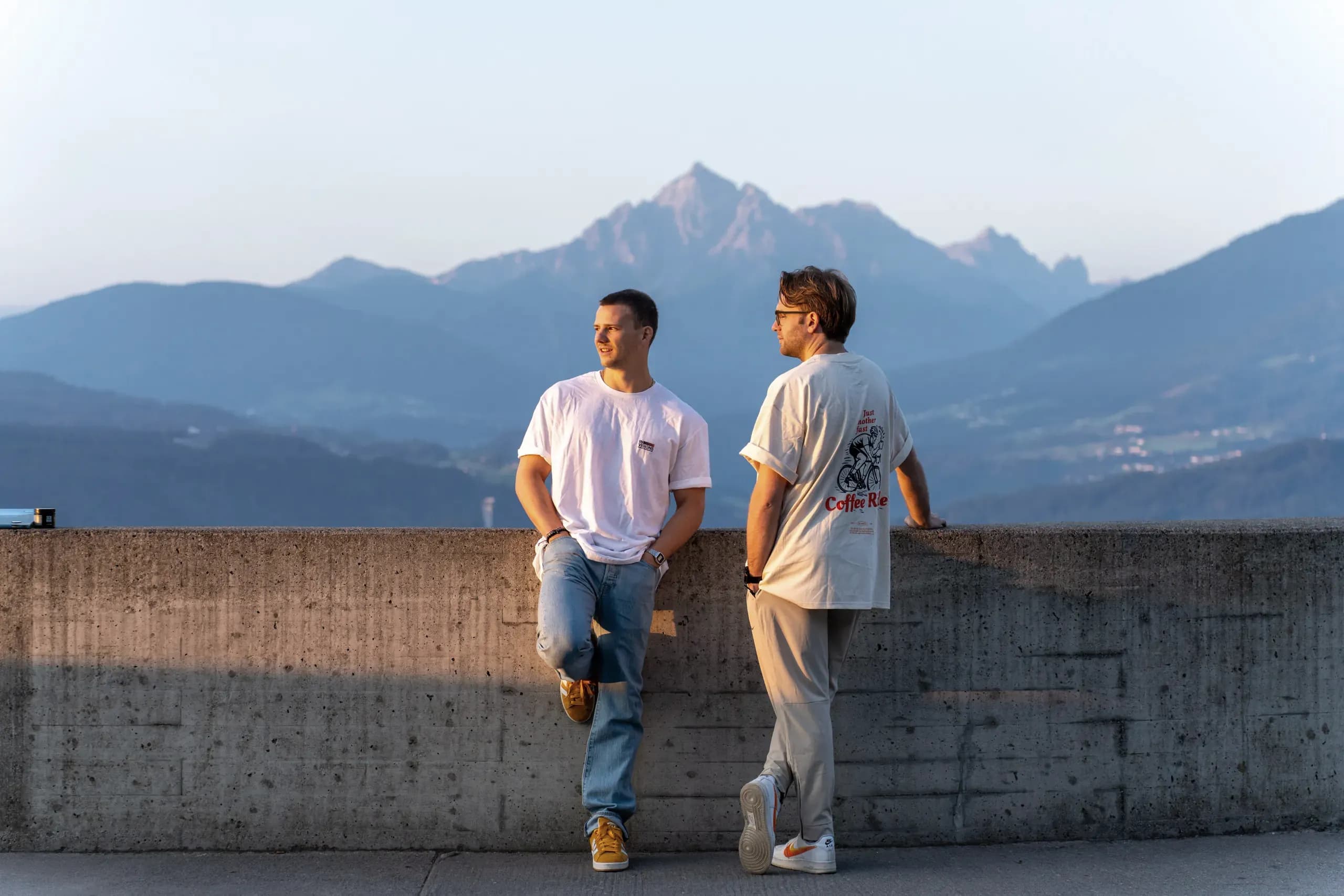 Zwei aus dem Team lehnen im Abendlicht an einer Mauer, dahinter ein Bergpanorama.