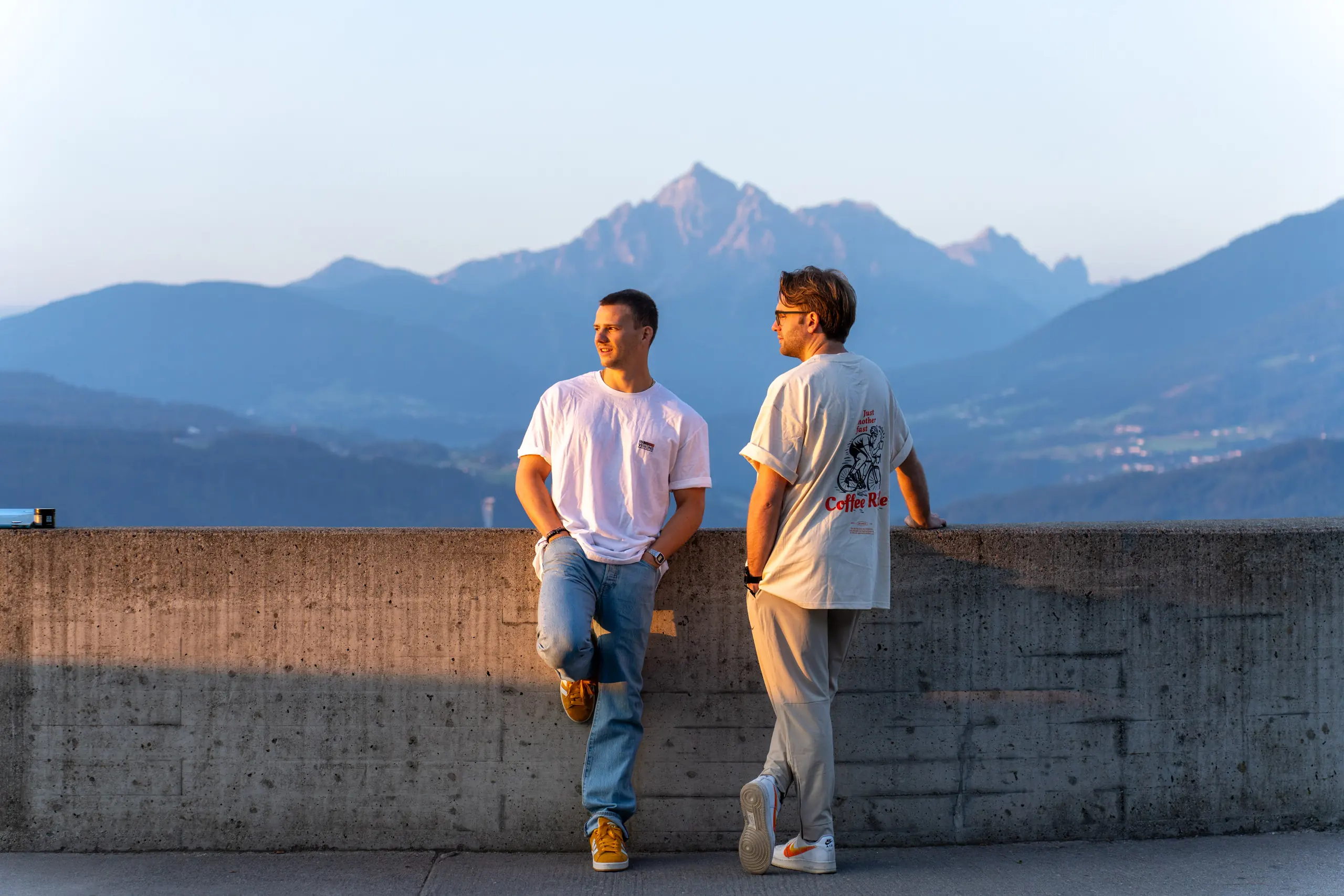 Zwei aus dem Team lehnen im Abendlicht an einer Mauer, dahinter ein Bergpanorama.