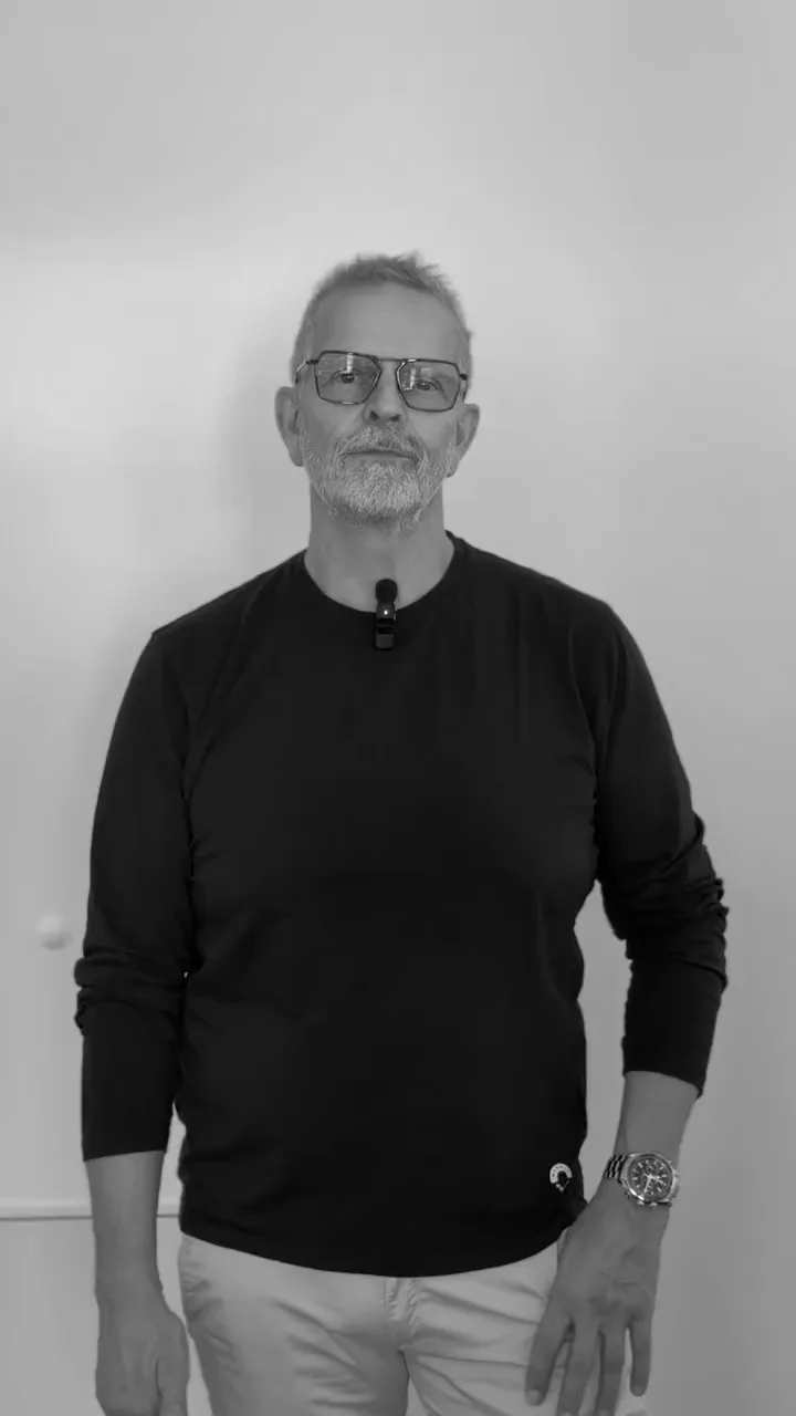 Standbild aus der Interviewreihe: ein Mann mit Brille in schwarzem Langarmshirt und Ansteckmikrofon steht in Schwarzweiß vor einer hellen Wand.