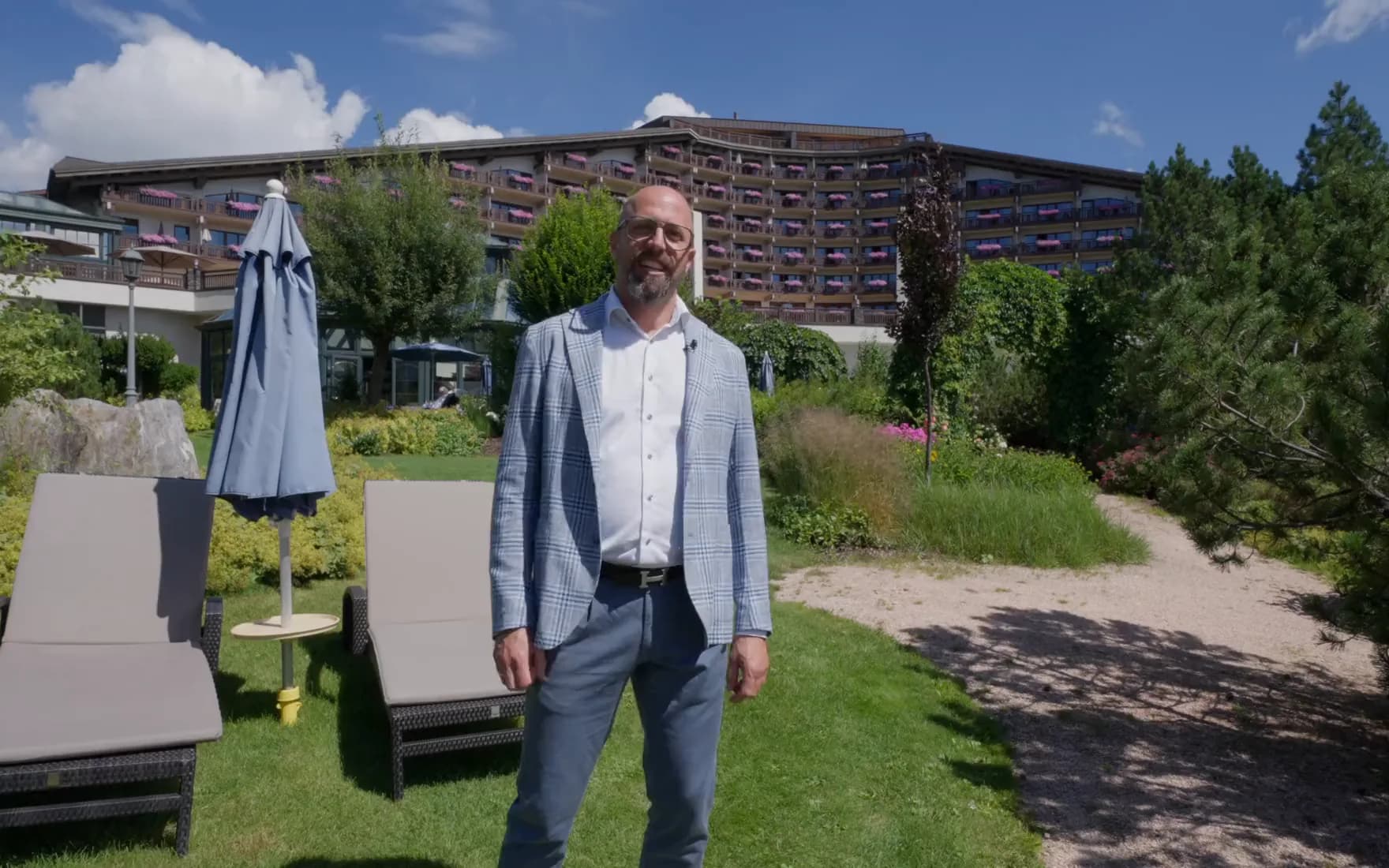 Der Film über das Interalpen Hotel: ein Mitarbeiter von AVsolutions im hellen Sakko geht vom Garten durch Konferenzbereich, Seminarraum, Suite, Bar und Almhütte und erklärt die Anlagen.