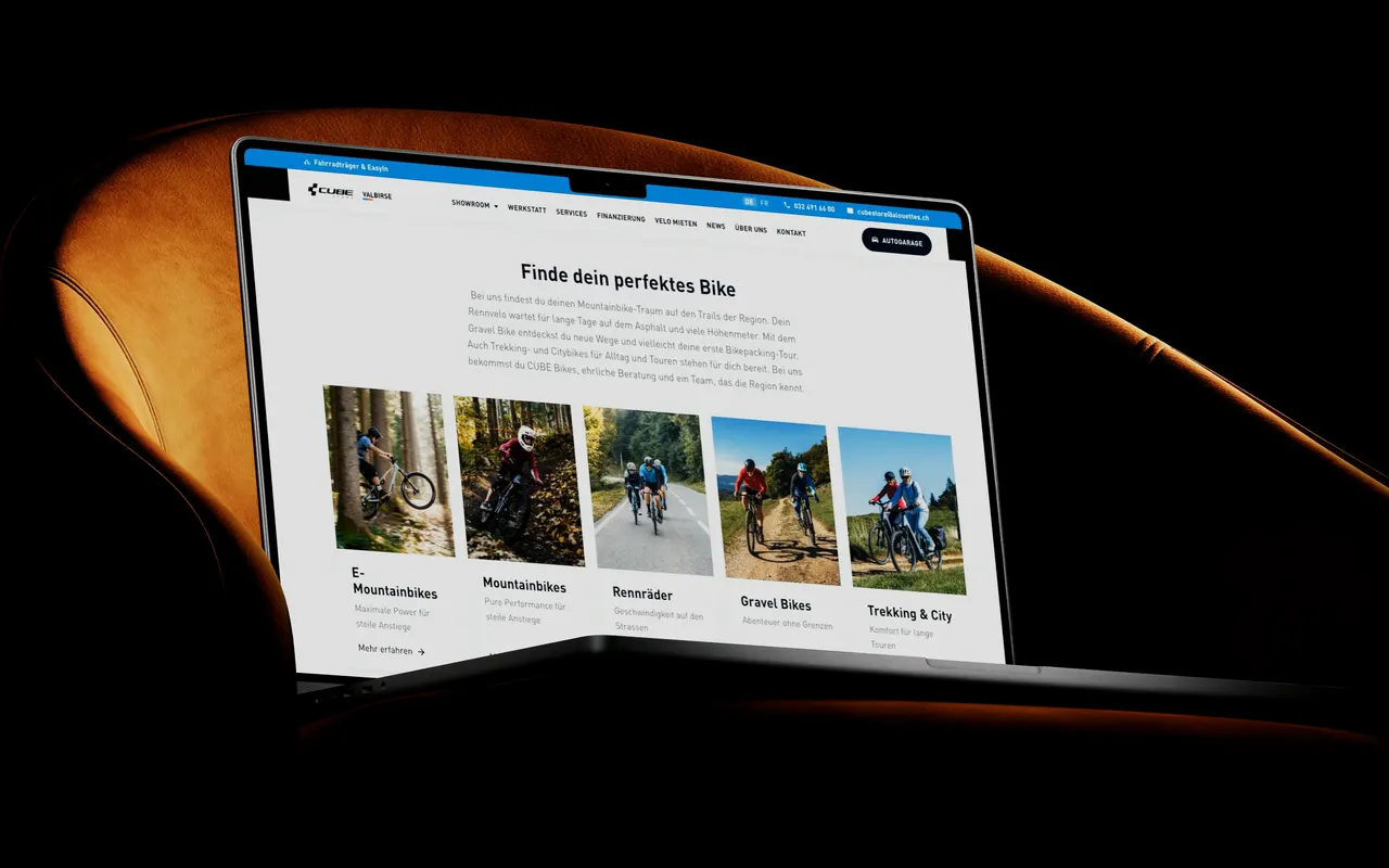 Die Startseite von cubestorevalbirse.ch auf einem MacBook vor einem orangefarbenen Sessel: das CUBE Modelljahr 2027, darunter die fünf Kategorien mit Bildern aus dem Jura und der Textteil über den Laden.