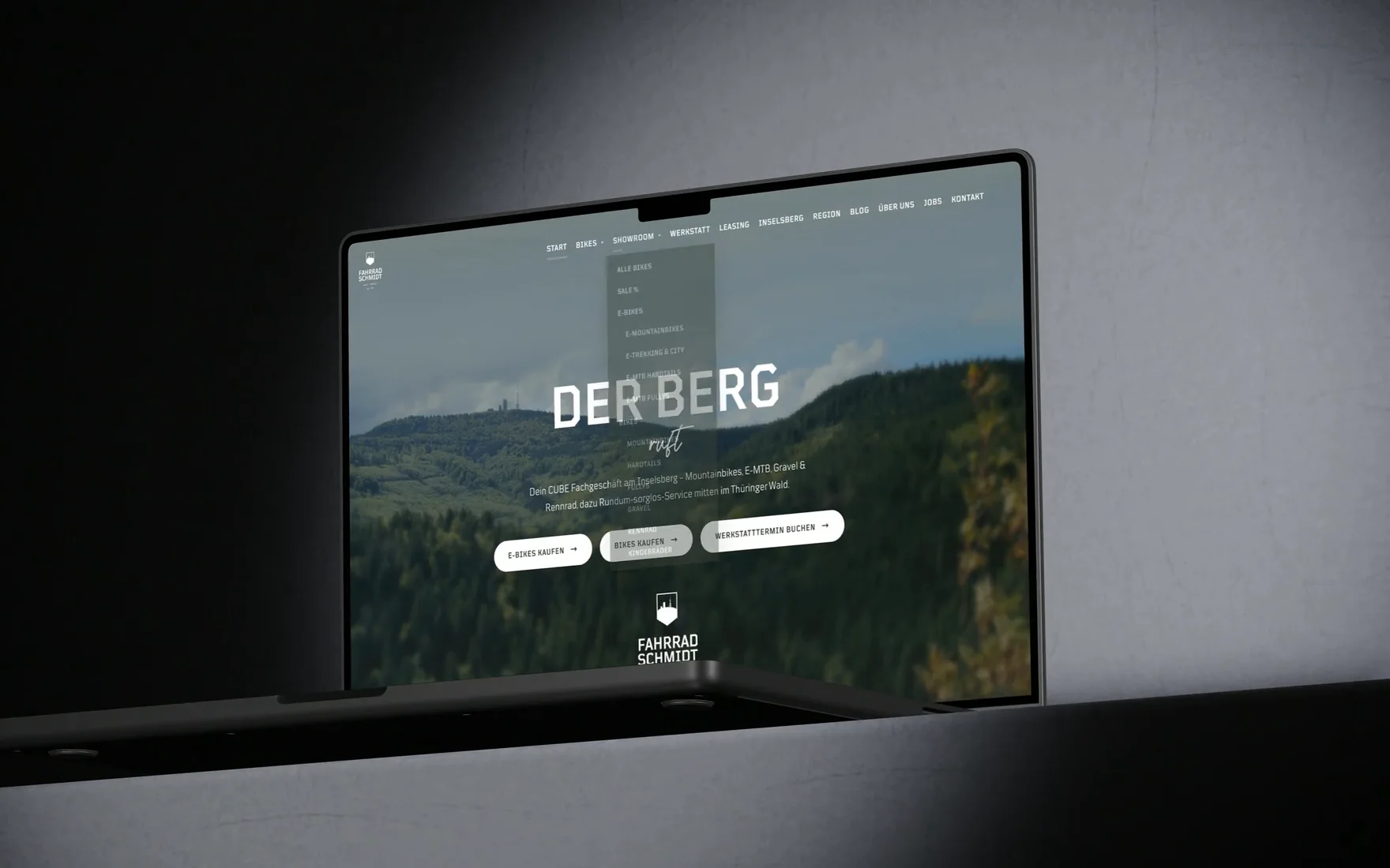Ein MacBook Pro öffnet sich in einem dunklen Raum und zeigt die Startseite von fahrrad-schmidt.de mit dem Titel Der Berg ruft, danach klappt das Showroom-Menü mit seinen Kategorien auf.