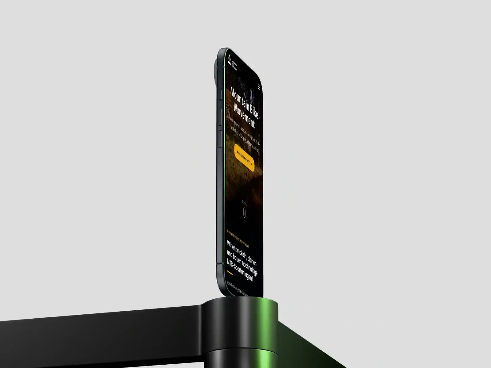 Ein iPhone auf einem Sockel vor hellgrauem Grund zeigt die mobile Startseite von mountainbikemovement.com mit dem Schriftzug Mountain Bike Movement über einem Waldtrail.