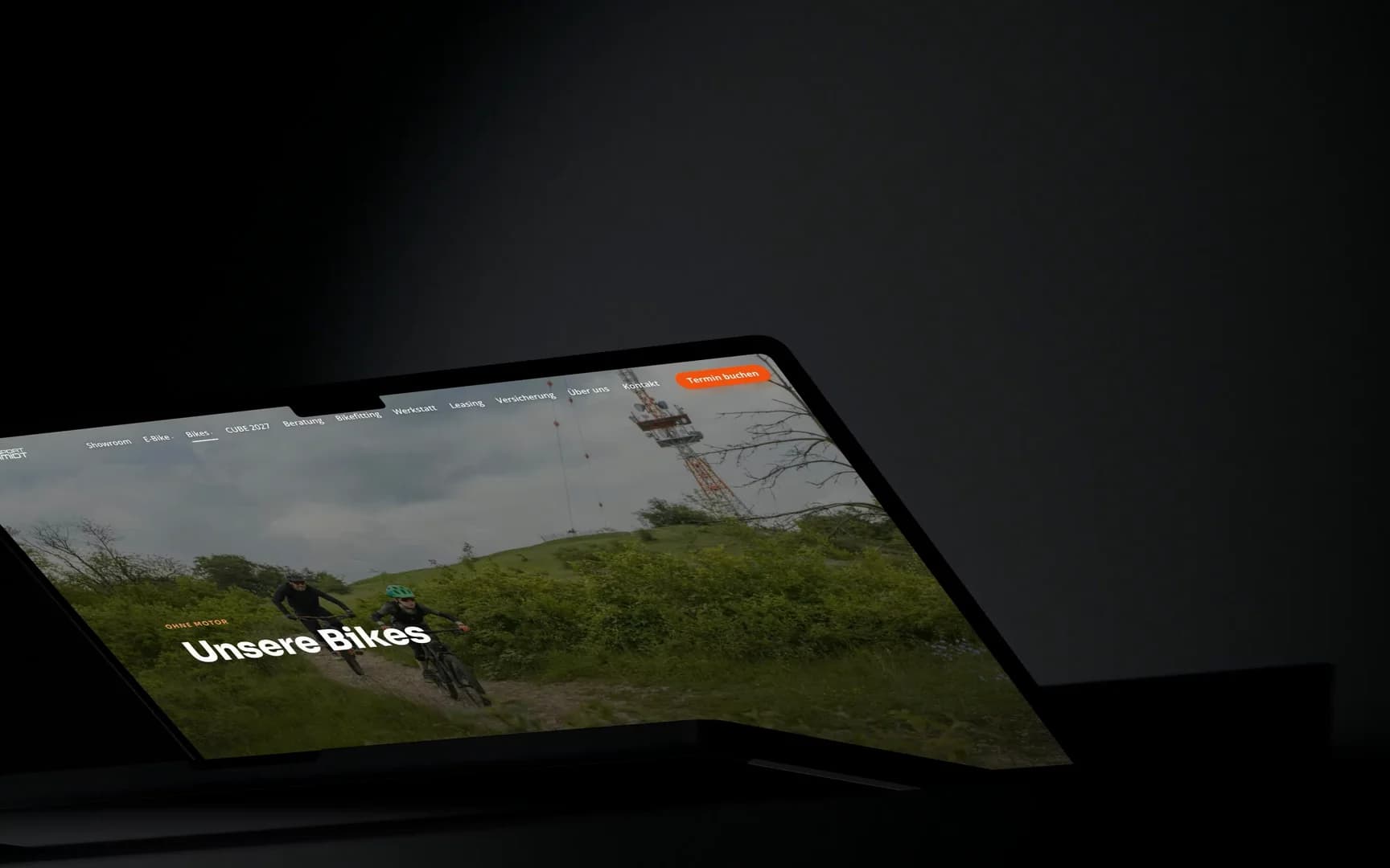 Ein MacBook im Lichtkegel an einer grauen Wand; auf dem Bildschirm läuft die Bikes-Seite von rad-sport-schmidt.de: der Shop mit seiner Filterleiste, der Filter Gravel und Rennrad, dann die Produktseite eines Cube Nuroad bis in den Seitenfuß.
