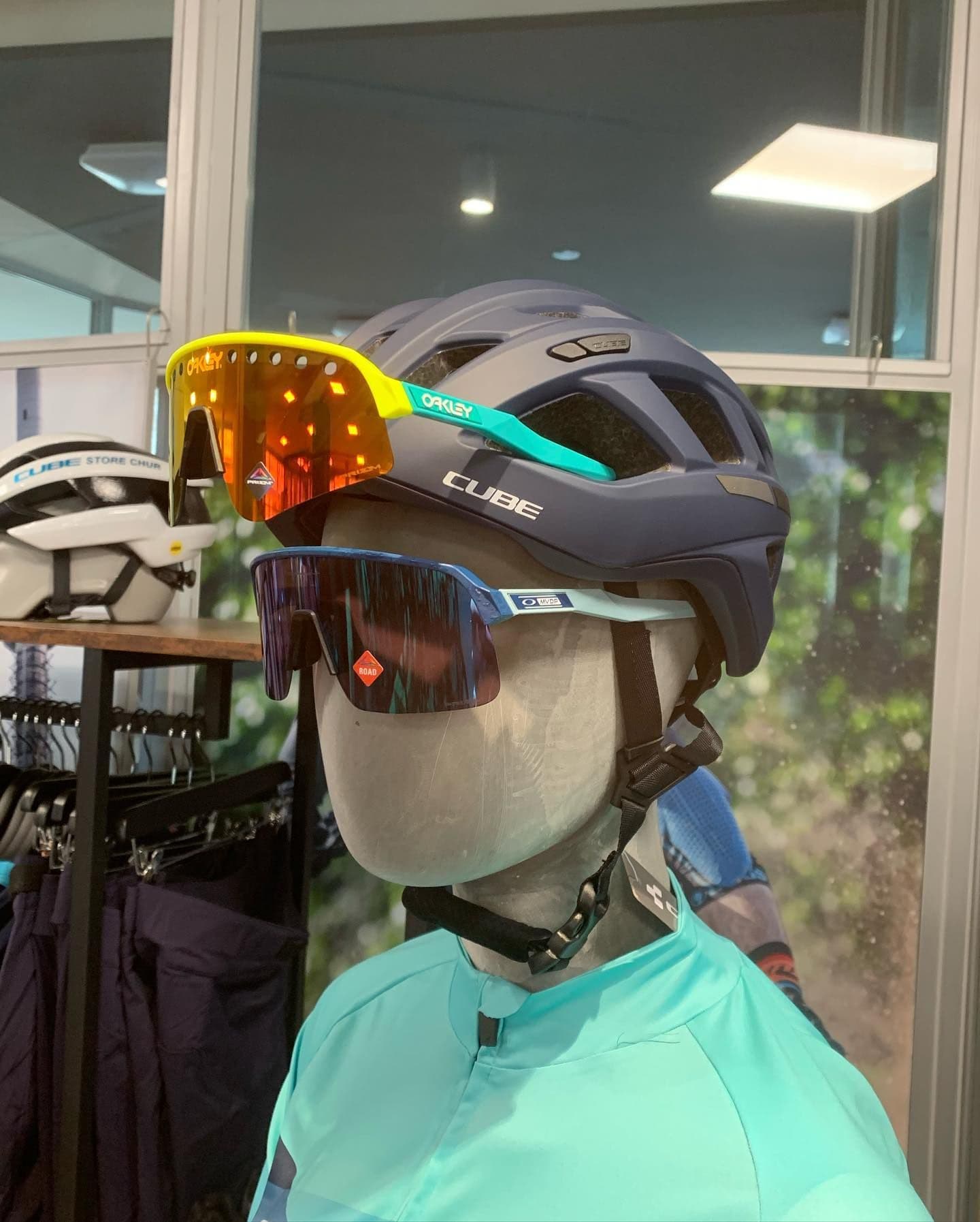 Eine Schaufensterpuppe im mintgrünen Trikot trägt einen dunkelblauen CUBE-Helm und zwei Brillen übereinander, im Regal dahinter ein weisser Helm mit dem Aufdruck des CUBE STORE CHUR.
