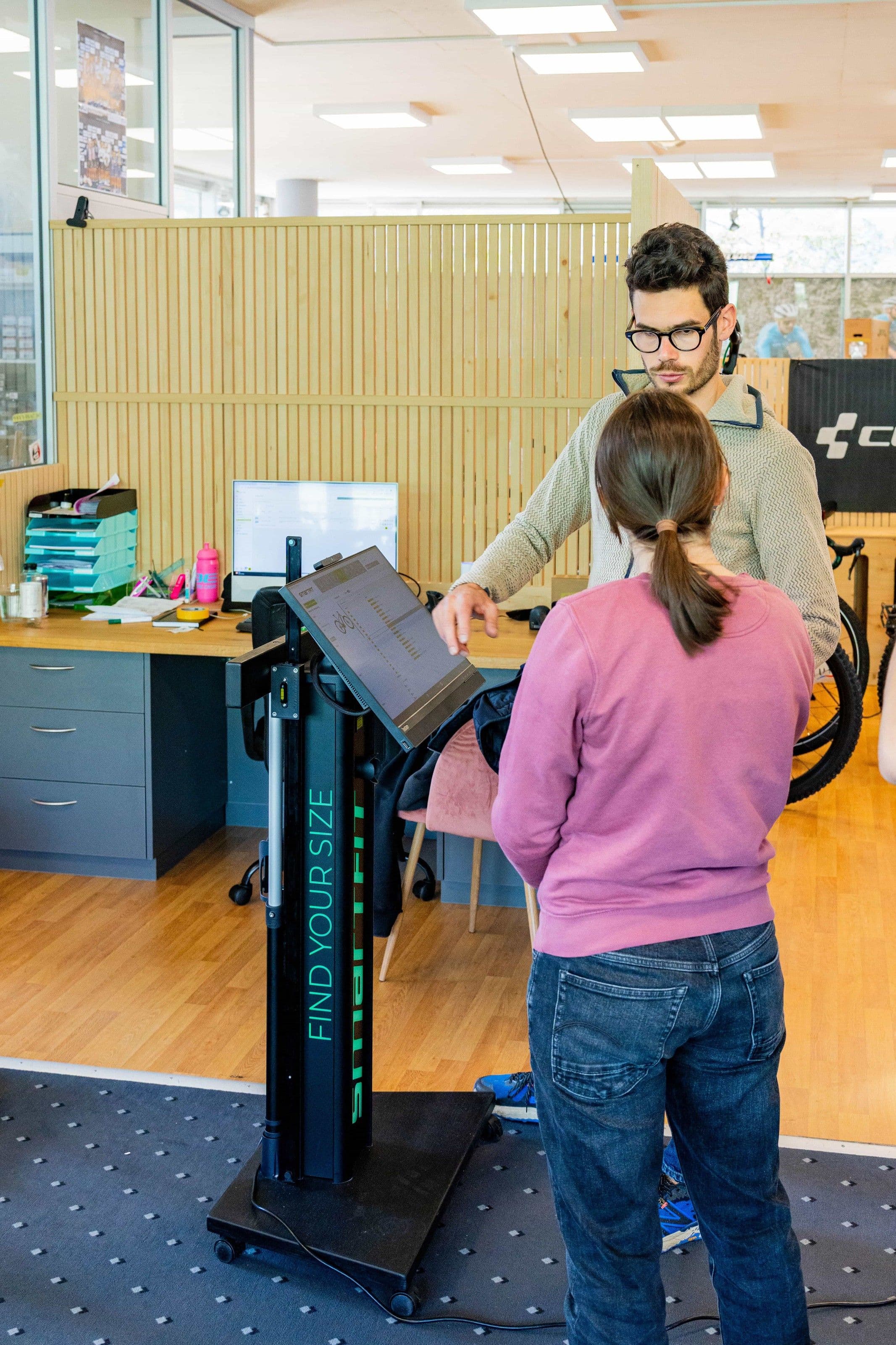 Ein Mitarbeiter des CUBE STORE CHUR steht hinter einer Kundin und zeigt ihr die Werte auf dem Bildschirm der Messsäule für die Rahmengrösse.