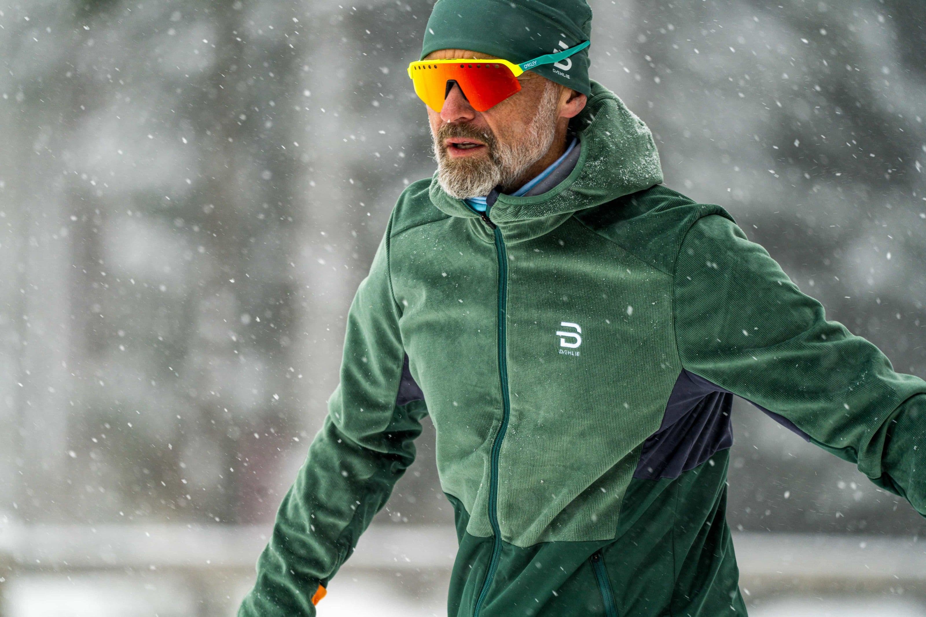 Nahaufnahme eines bärtigen Langläufers in grünem Anzug mit Mütze und orange verspiegelter Brille im Skating-Schritt, dichter Schneefall vor weissem Hintergrund.