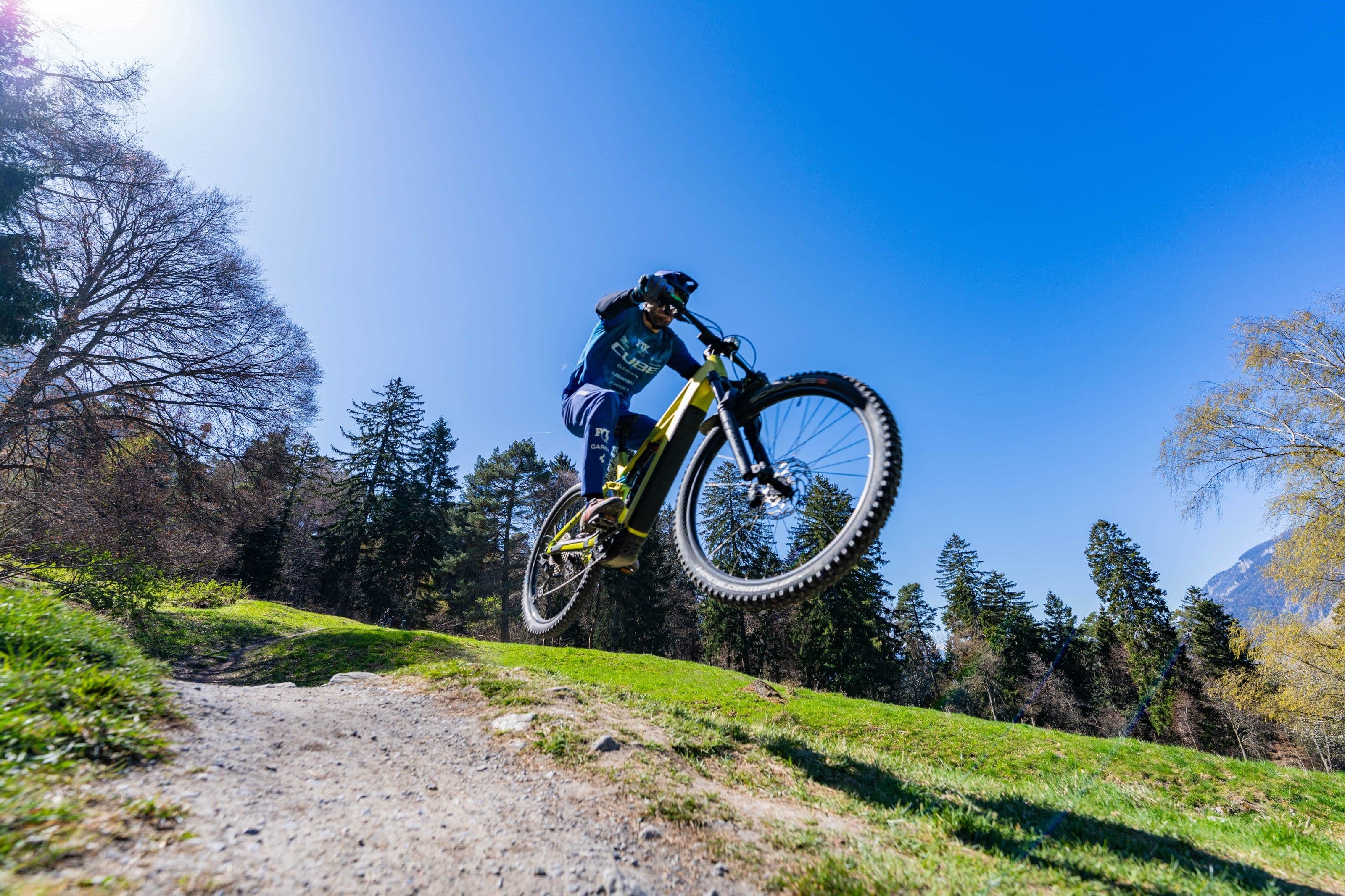 Aus der Froschperspektive: ein Fahrer springt mit einem gelben CUBE E-Mountainbike über eine Erdkante, beide Räder in der Luft vor tiefblauem Himmel.