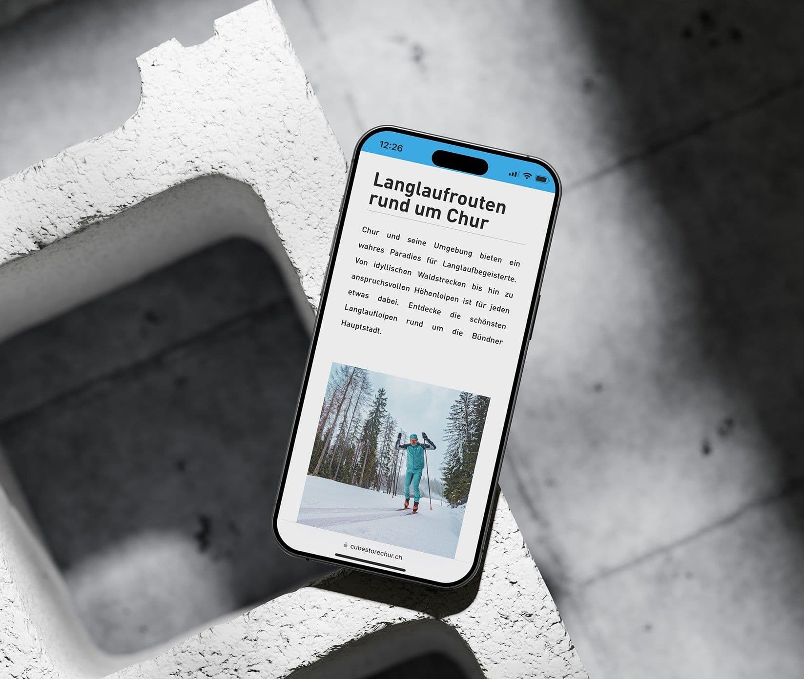 Ein iPhone liegt schräg auf weissen Betonformteilen, auf dem Bildschirm ein Ratgeber des Stores über Langlauf rund um Chur mit dem Foto einer Langläuferin im verschneiten Wald, unten die Adresszeile cubestorechur.ch.