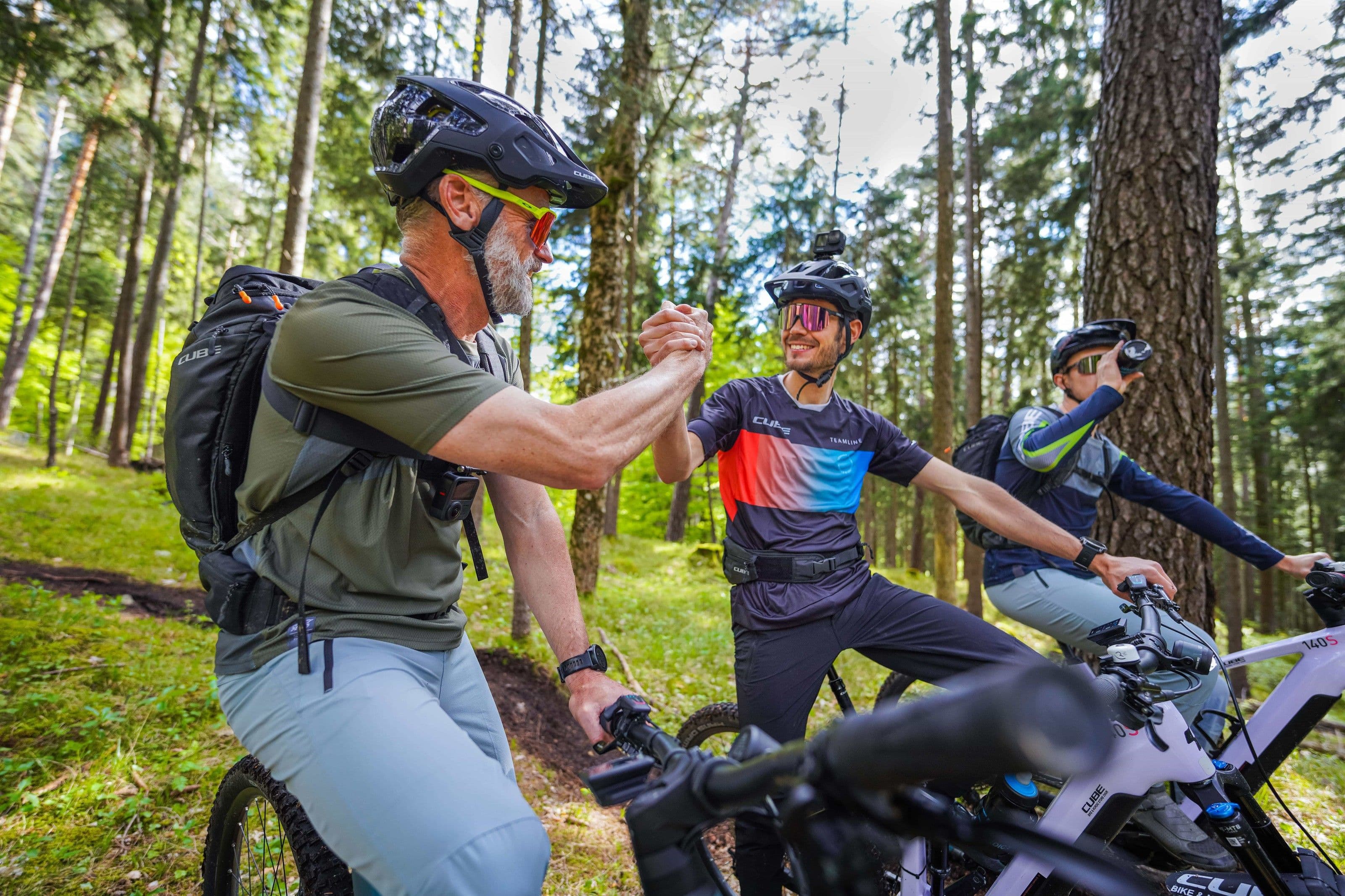 Zwei Mountainbiker halten im lichten Nadelwald an und schlagen über dem Lenker lachend die Hände ineinander, ein dritter Fahrer steht dahinter.