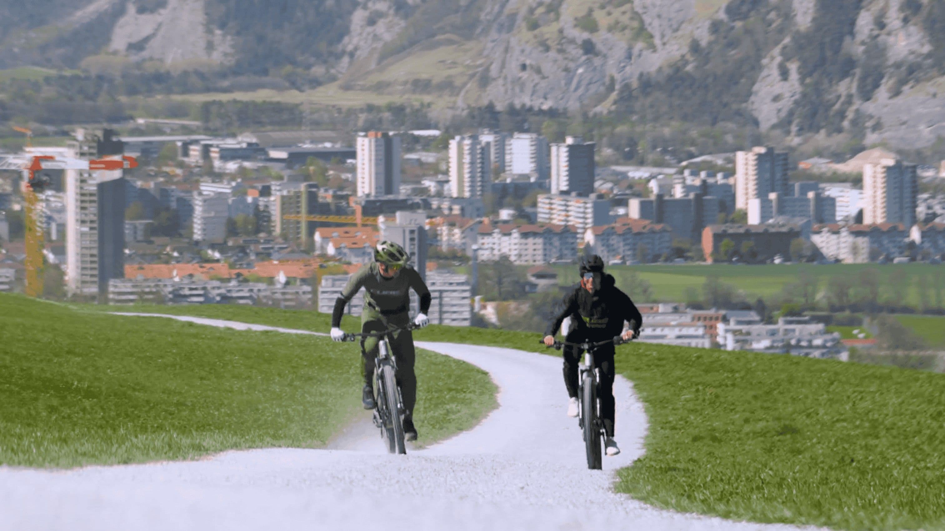 Zwei Mountainbiker kommen einen hellen Kiesweg über einer Wiese auf die Kamera zu, dahinter die Hochhäuser von Chur und die Felswand des Calanda.