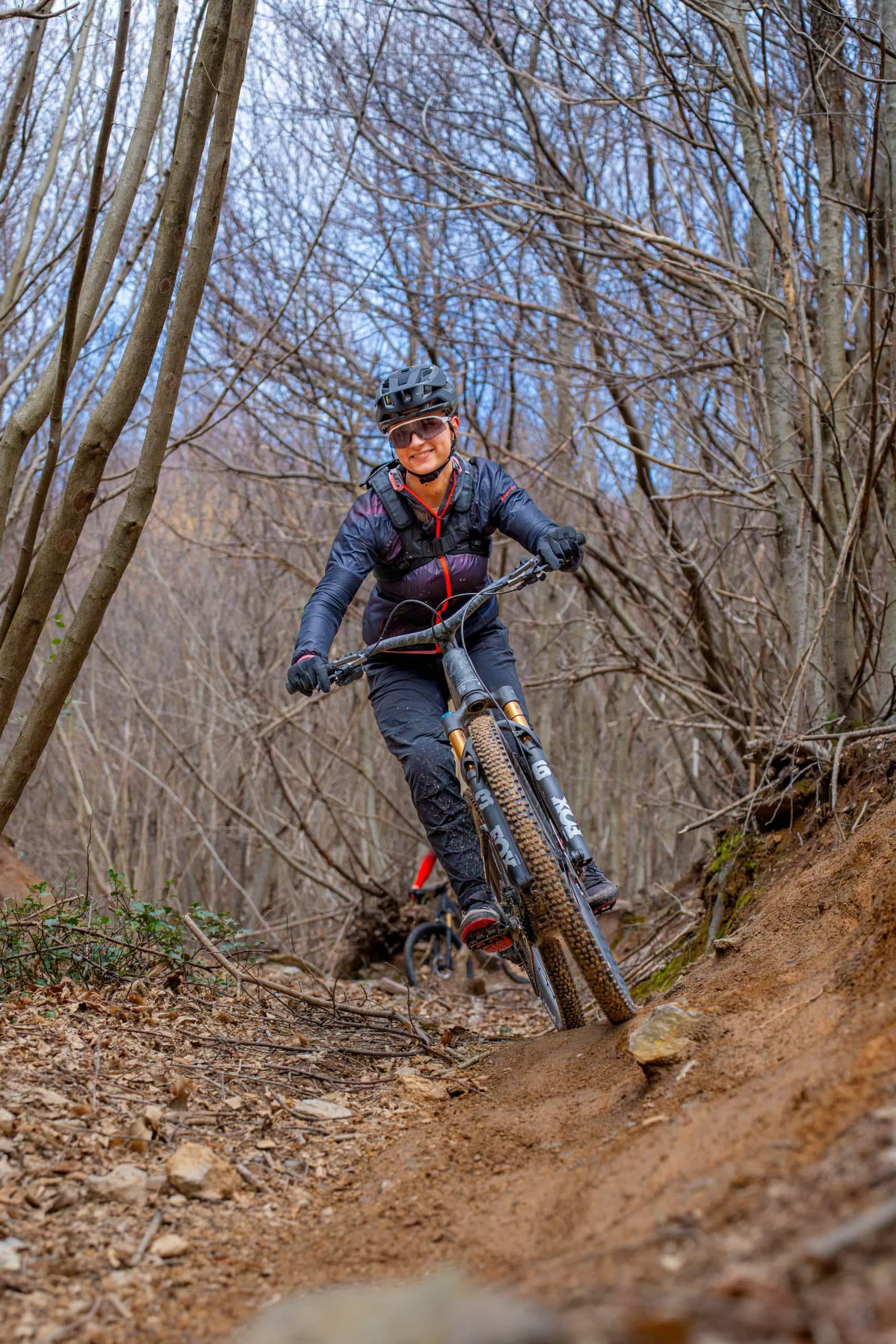 Eine lachende Mountainbikerin in dunkelblauer Jacke fährt eine ausgewaschene Erdrinne im kahlen Laubwald hinunter.