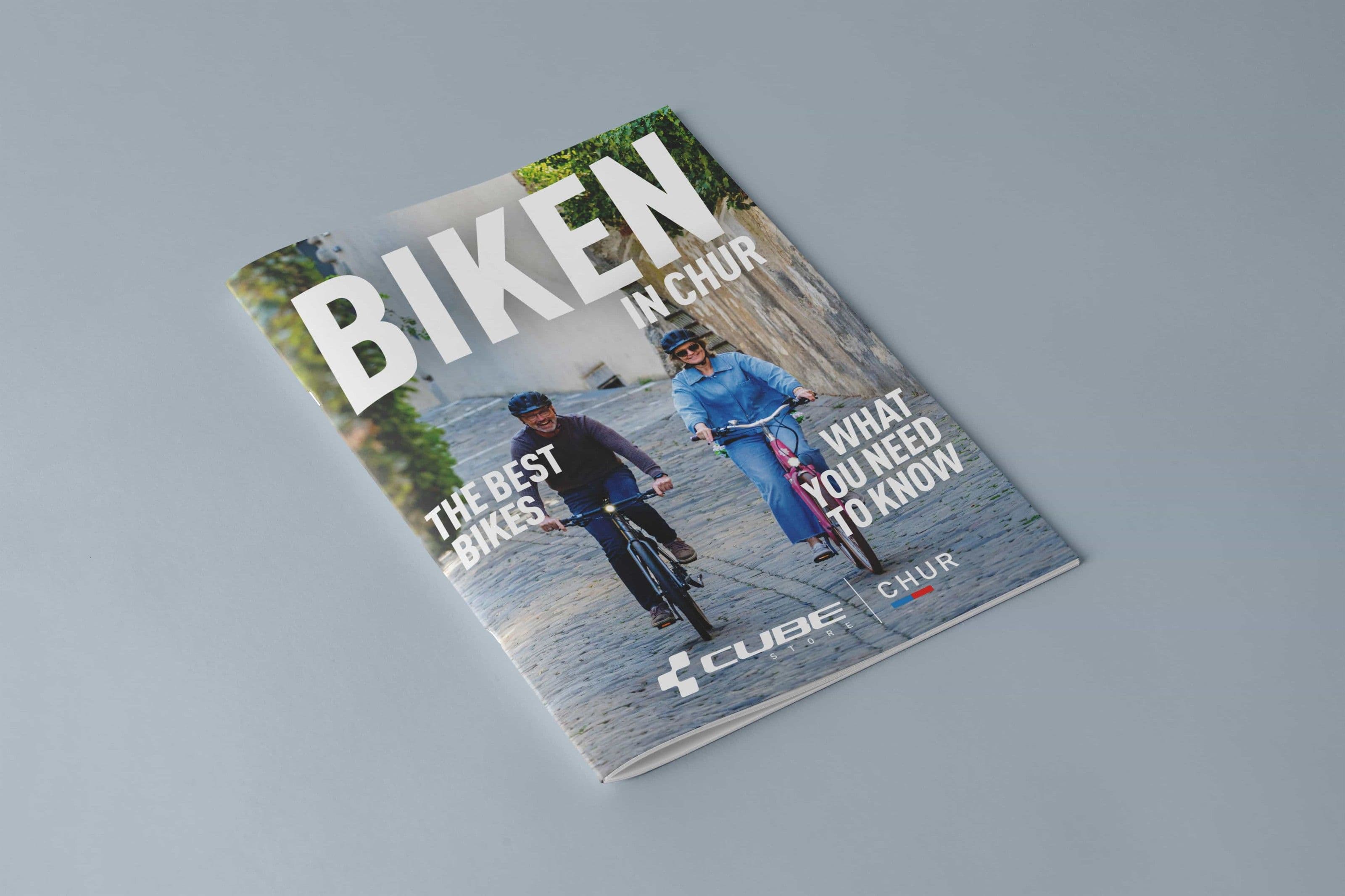 Das Magazin BIKEN IN CHUR liegt geschlossen auf hellgrauem Grund, auf dem Titel zwei Freizeitradler auf Kopfsteinpflaster und die Doppelmarke CUBE STORE Chur.