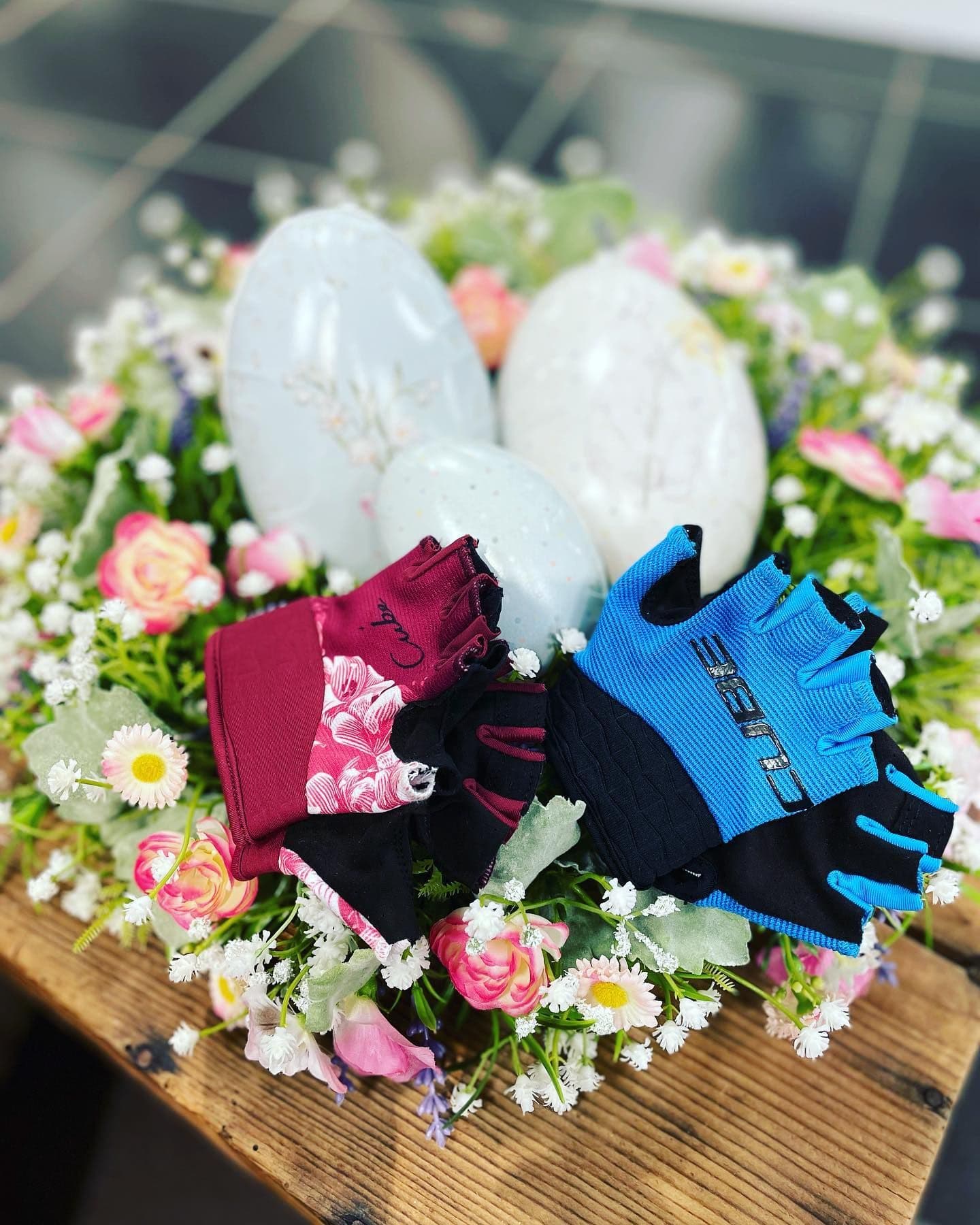 Zwei Paar CUBE-Radhandschuhe, eines weinrot mit Blumenmuster, eines hellblau, liegen in einem Osterarrangement aus Kunstblumen und drei weissen Eiern auf einer alten Holzkiste.
