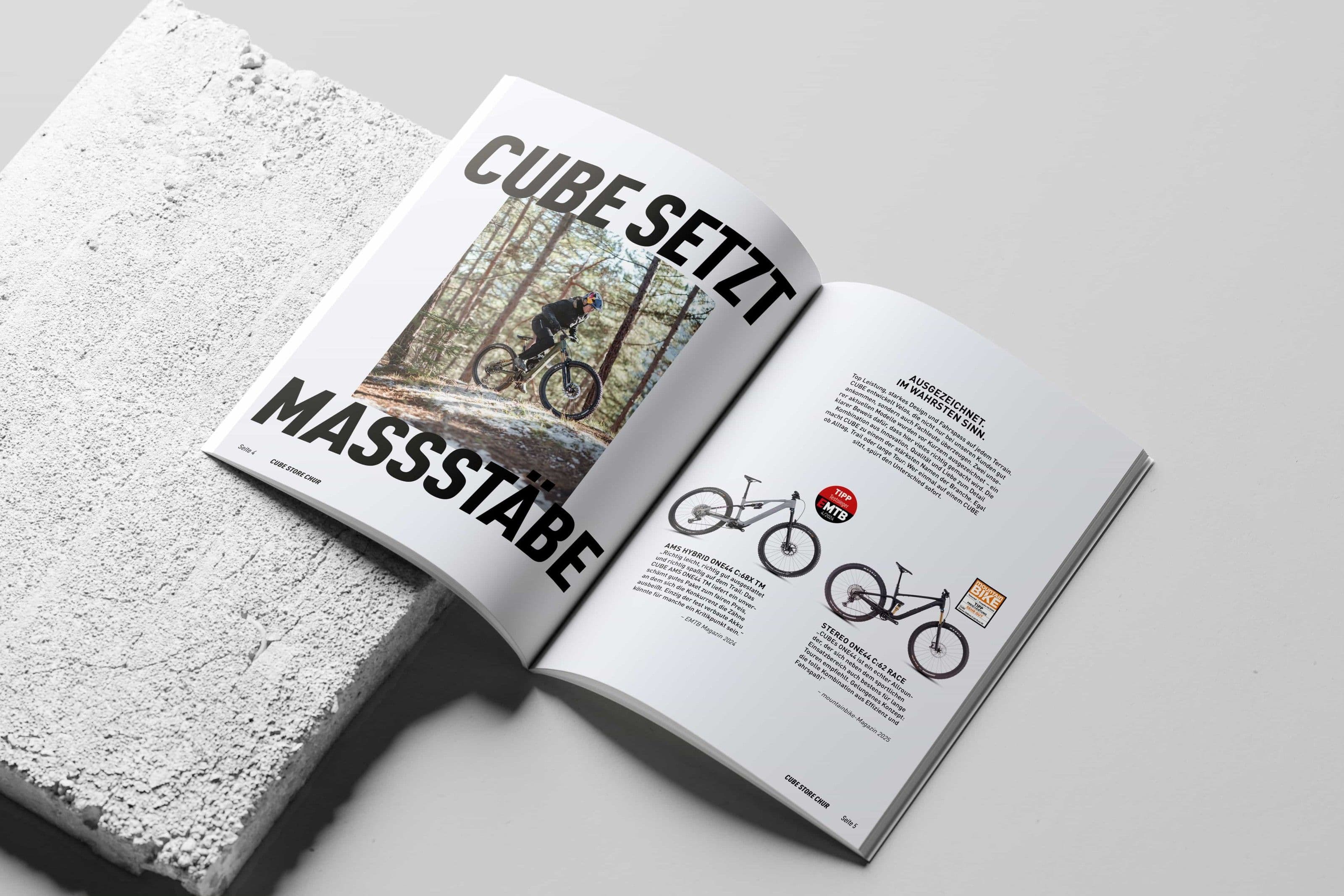 Ein aufgeschlagenes Magazin auf einem weissen Betonblock: links die Schlagzeile CUBE SETZT MASSSTÄBE über einem Waldfoto mit Mountainbiker, rechts eine Textspalte mit zwei ausgezeichneten Bikes.