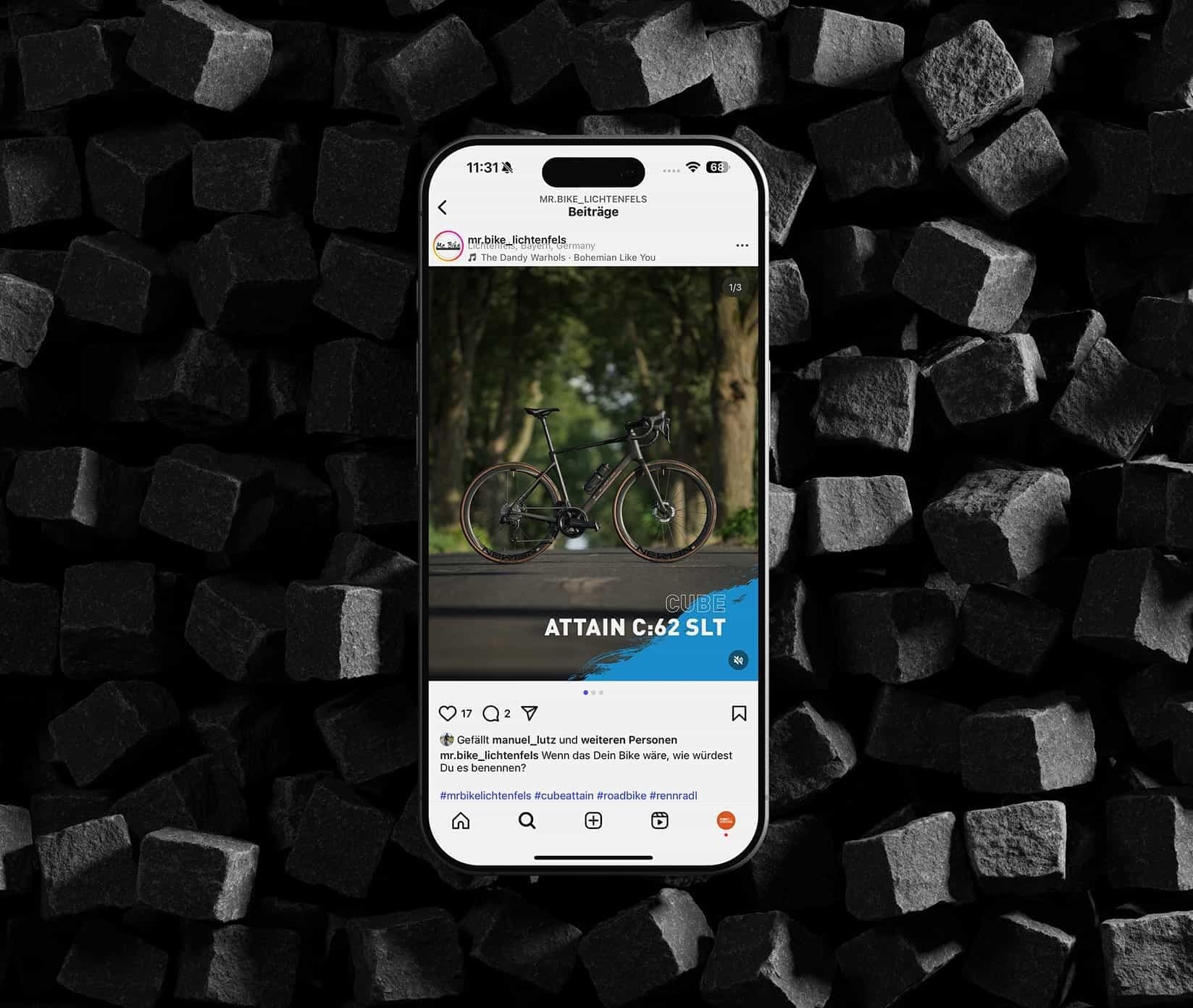 Ein Smartphone auf schwarzen Holzwürfeln zeigt einen Instagram-Beitrag von mr.bike_lichtenfels: ein schwarzes CUBE Rennrad auf einem Waldweg, darunter die Zeile Attain C:62 SLT.