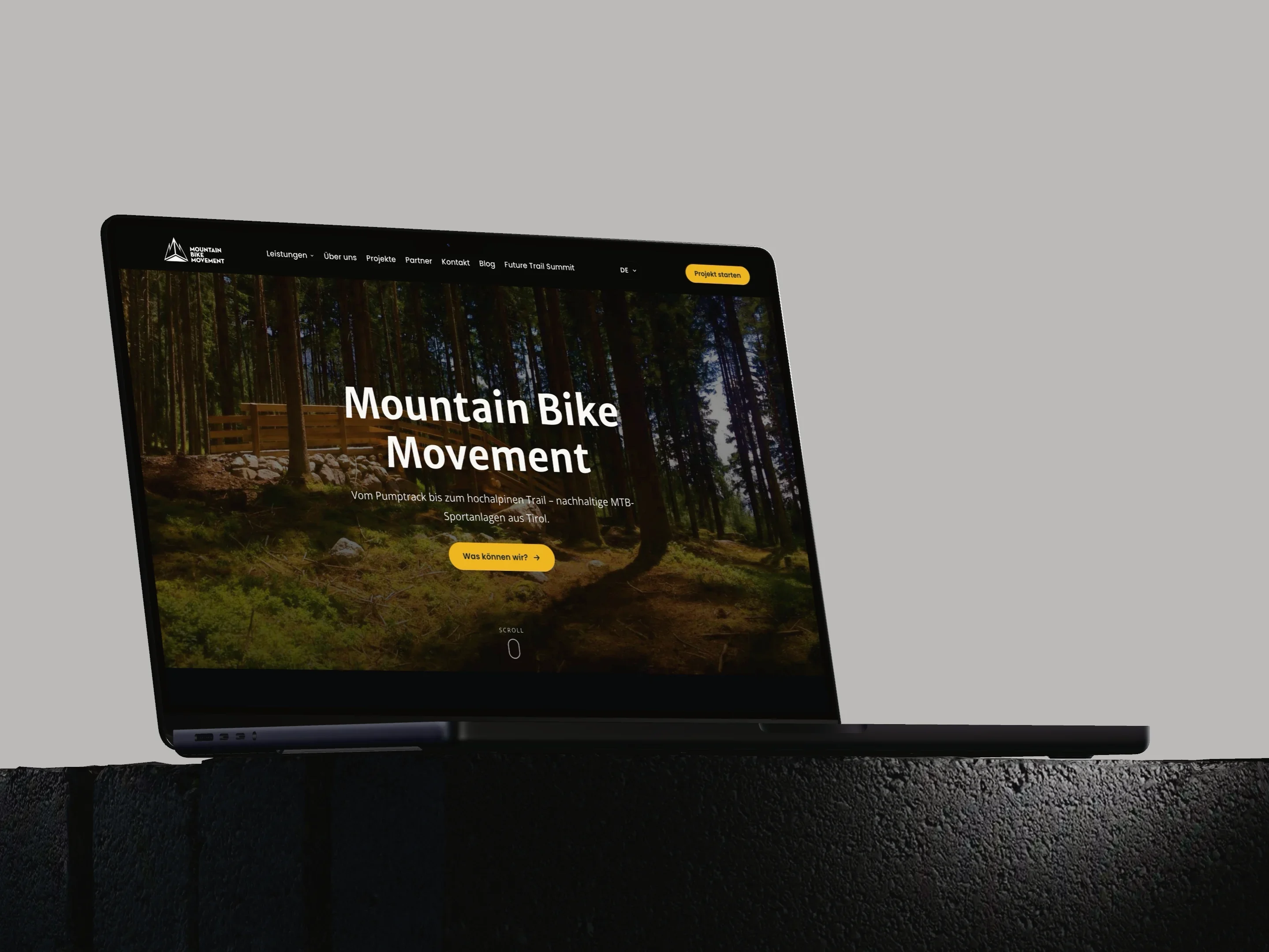 Mountain Bike Movement: Ein MacBook Pro auf einem dunklen Steinsockel zeigt die Startseite von mountainbikemovement.com mit einem Waldtrail.