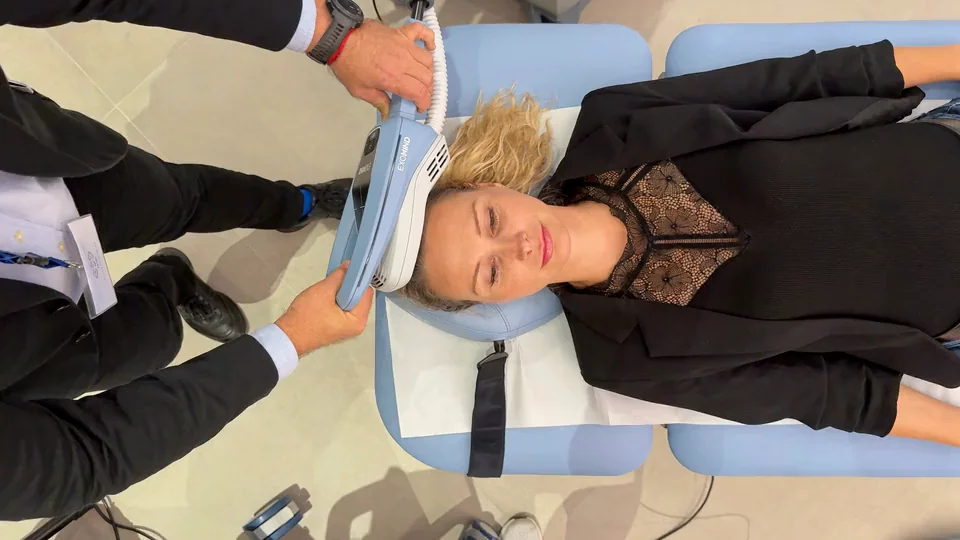 Behandlung von oben: eine Patientin liegt mit geschlossenen Augen auf einer hellblauen Liege, ein Arzt im dunklen Sakko hält ein Behandlungsgerät an ihre Schläfe.