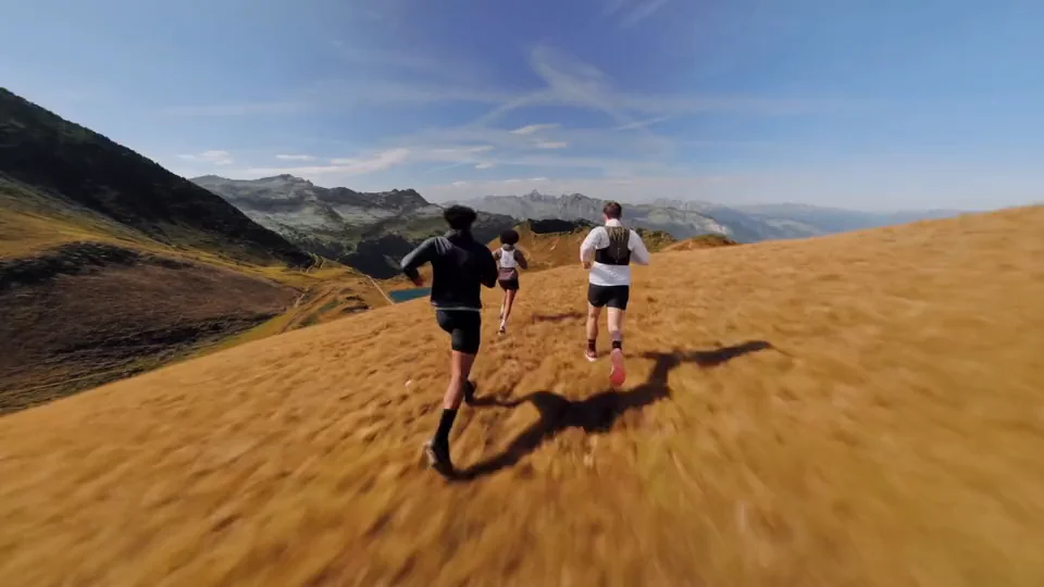 Drei Trailrunner laufen über einen Grasrücken bergab, dahinter ein Bergsee und eine Gebirgskette unter blauem Himmel.