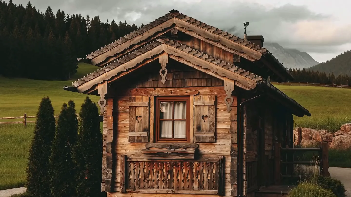 Kleines Holzchalet mit geschnitzten Fensterläden und Balkon auf einer Bergwiese, dahinter Nadelwald und Berge unter Wolken.