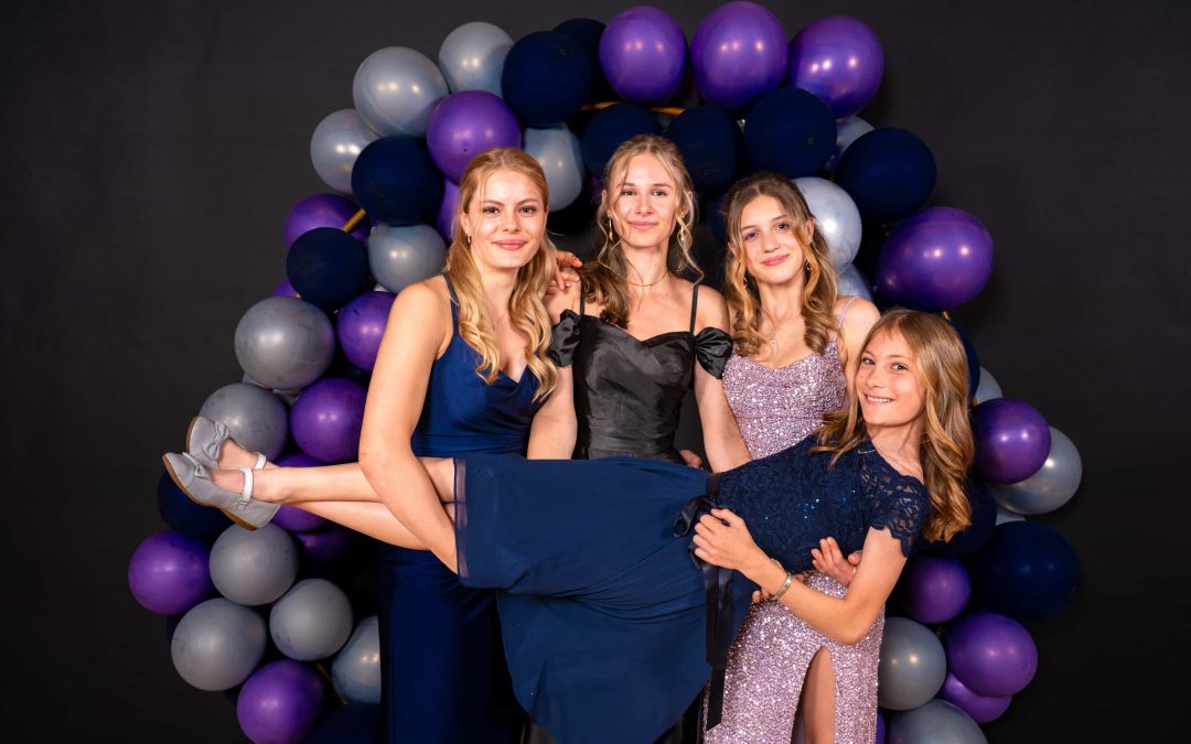 Vier junge Frauen in Abendkleidern posieren bei Invisions vor einem Ballonbogen in Lila, Blau und Silber. Drei stehen lächelnd da und halten die vierte hoch, die grinsend und waagerecht liegt – ein lustiges Foto, perfekt für Marketing-Erinnerungen.