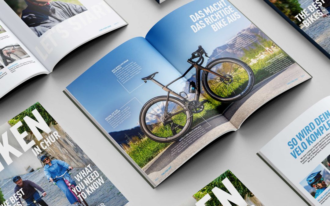 Eine Sammlung von offenen und geschlossenen Radsportmagazinen auf grauem Untergrund mit Bildern von Radfahrern und Fahrrädern – die ideale Inspiration für jede Marketingagentur, die sich auf Sportmarketing spezialisiert hat. Ein offenes Magazin zeigt ein Rennrad auf einem malerischen Weg mit deutschem Text.