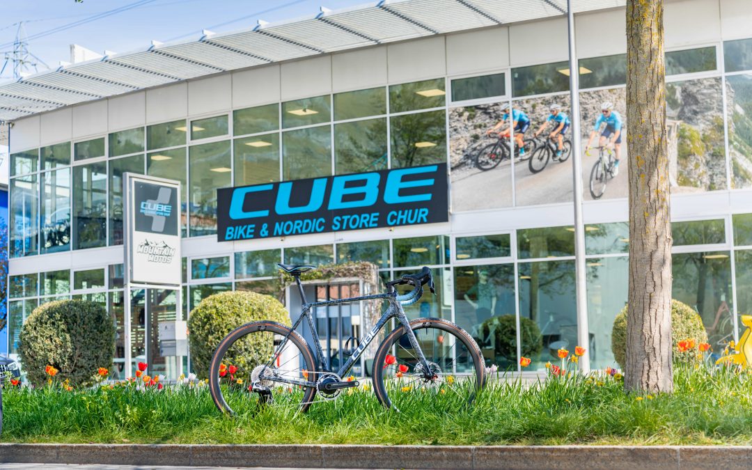 Ein schickes Rennrad steht vor dem Cube Bike & Nordic Store Chur, einem modernen Gebäude mit Glasfront – ein beeindruckendes Schaufenster für Sportmarketing mit Radfahrerfotos, leuchtendem Grün, Blumen und einem Baum, der die Szenerie verschönert.